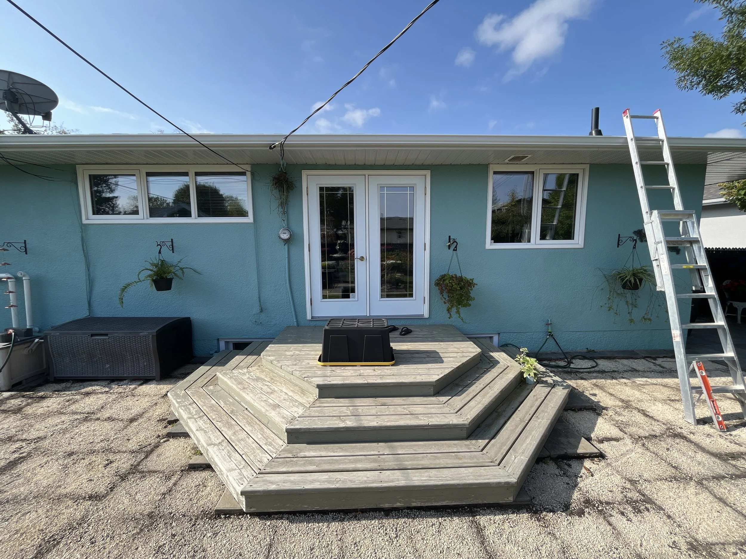 Backyard deck area in progress with wooden stairs leading up to a turquoise house, ladders, potted plants, and garden supplies visible.