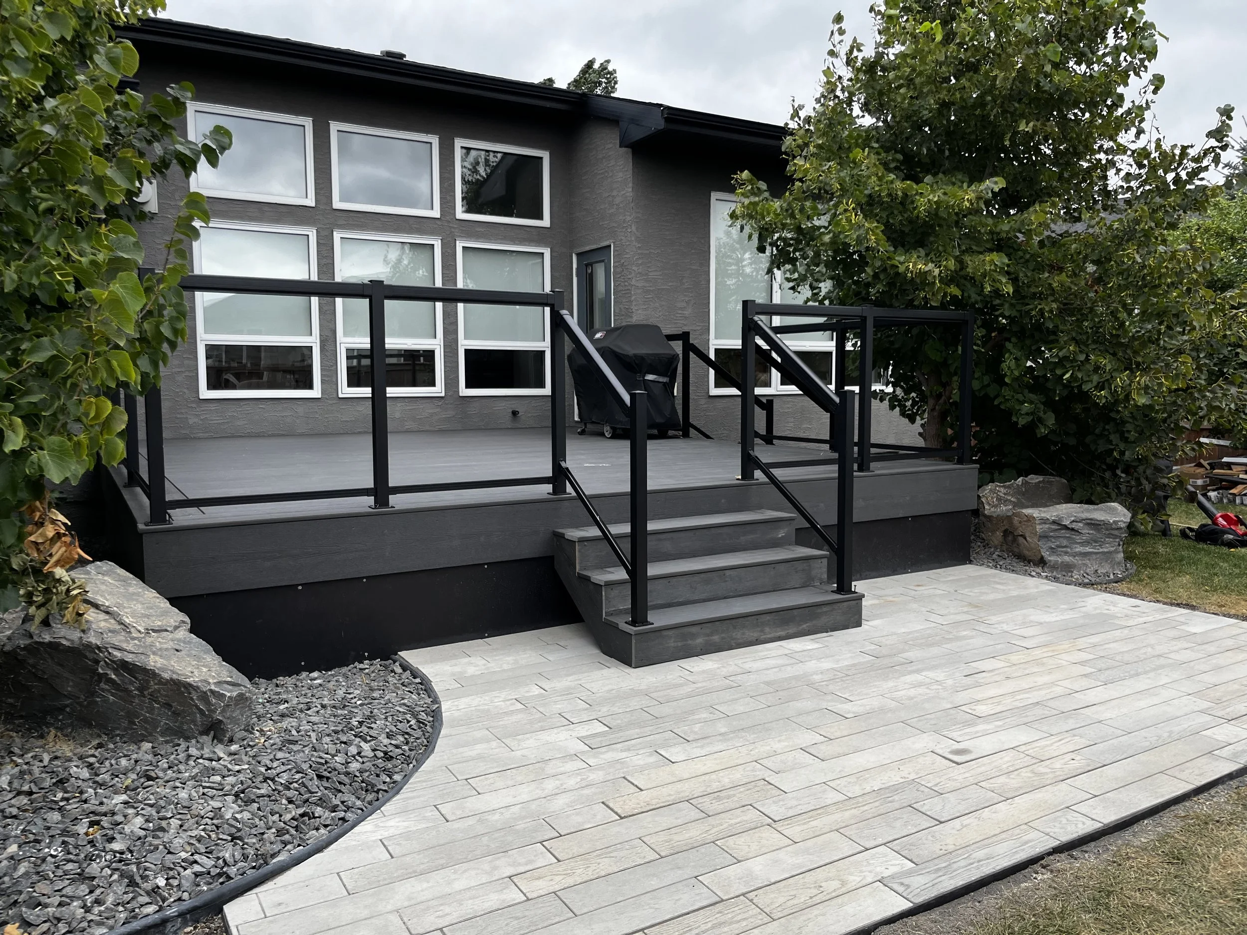 Gray house with large windows, black railing porch, and stairs leading up to it, surrounded by large rocks, trees, and a paved patio.