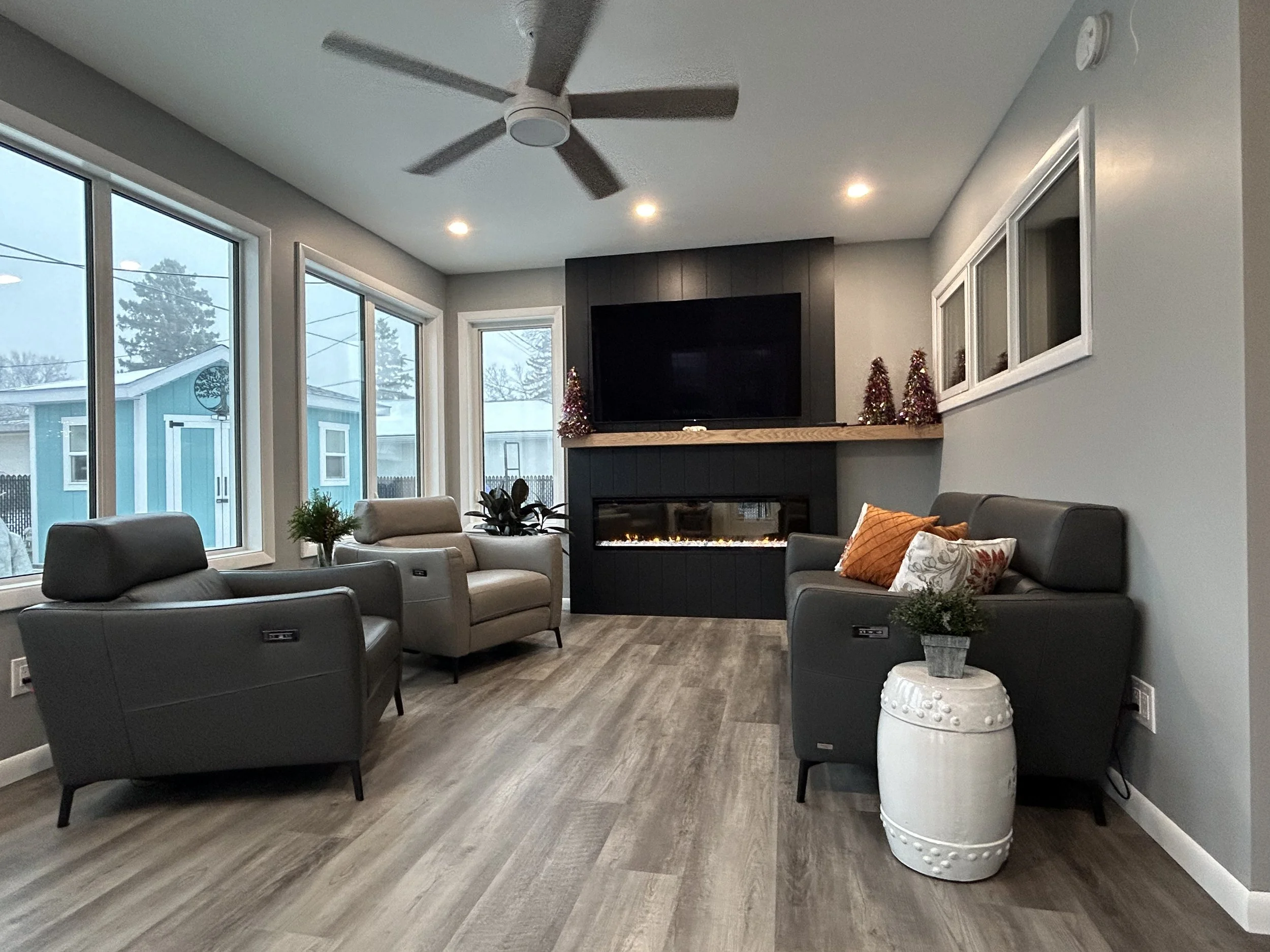 Living room with gray sofas, a fireplace, a TV above, large windows on left, a ceiling fan, hardwood floor, holiday decorations, and pillows on the sofas.
