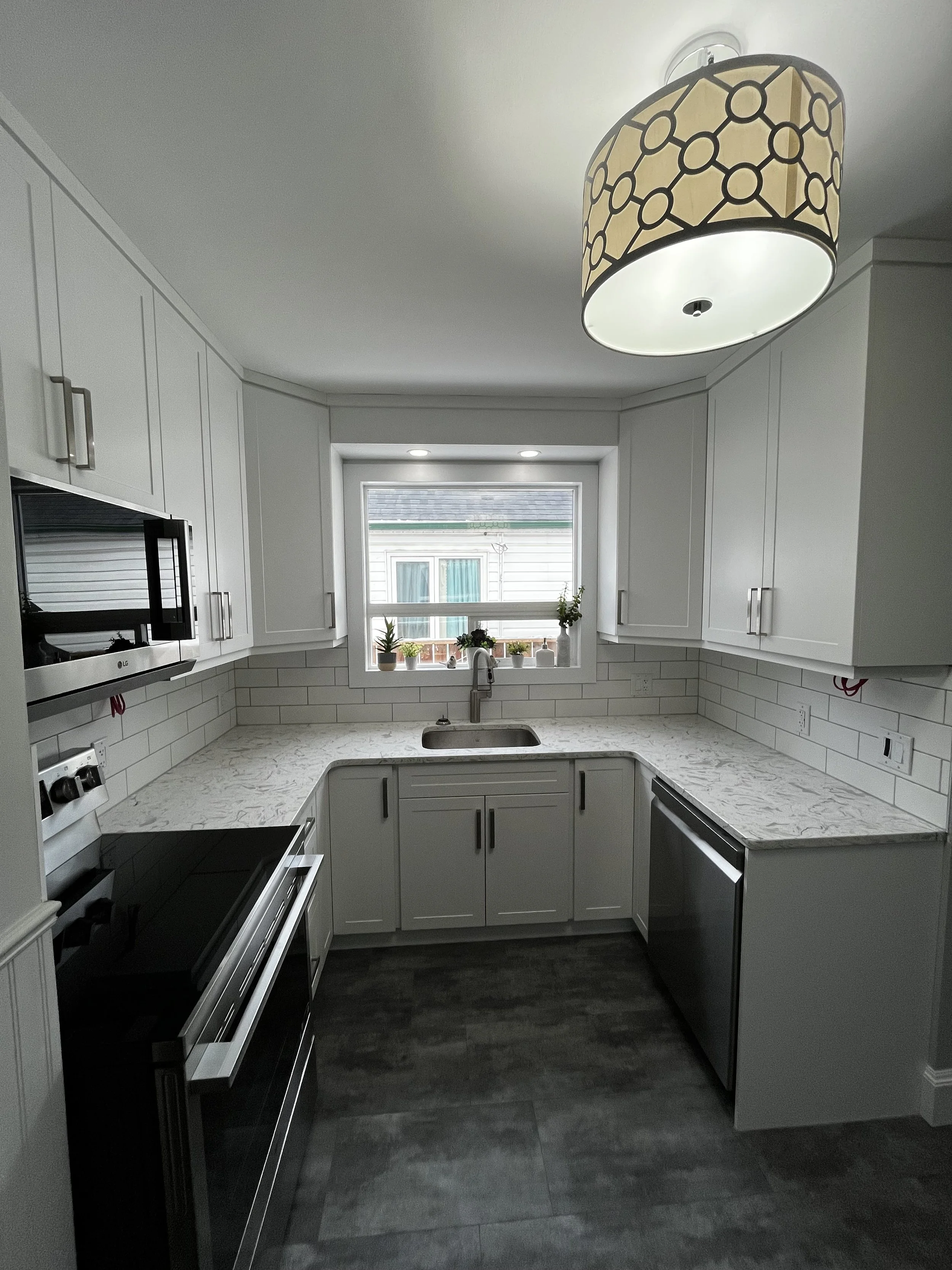 Modern kitchen with white cabinetry, marble countertops, stainless steel appliances, a small window above the sink with plants, gray flooring, and a decorative ceiling light fixture.