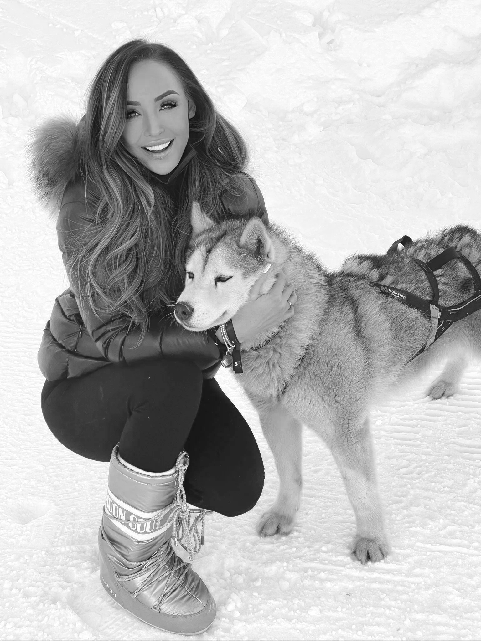 A woman in winter clothing kneels on snow, smiling, holding a Siberian Husky dog.