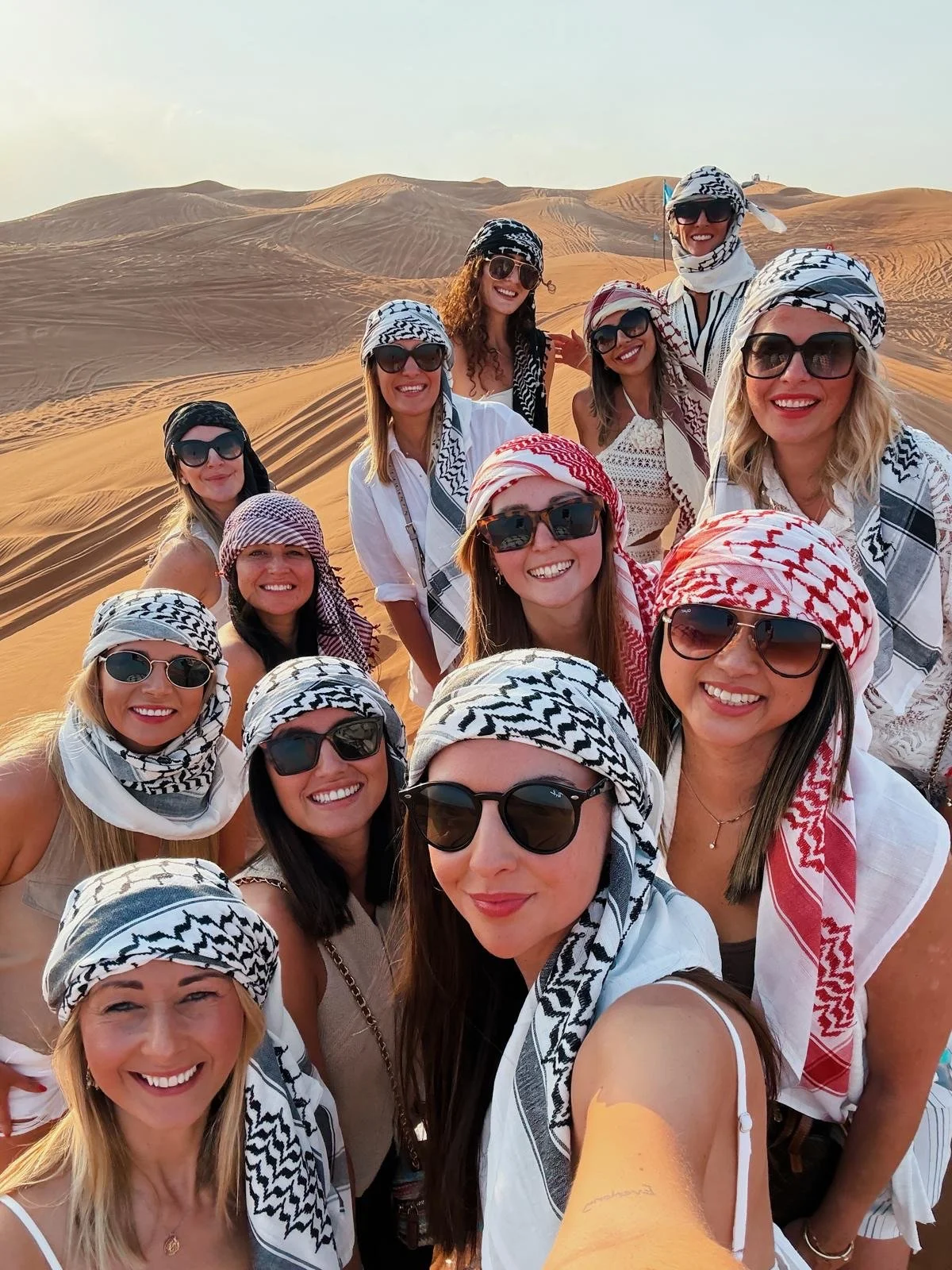 Group of women wearing sunglasses and traditional keffiyeh scarves taking a selfie in the desert with sand dunes in the background.