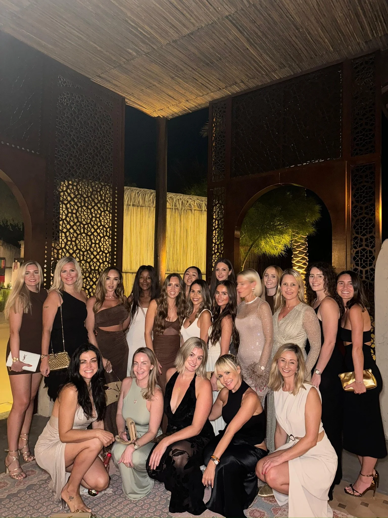 A large group of women dressed in formal and evening attire posing together outdoors at night.