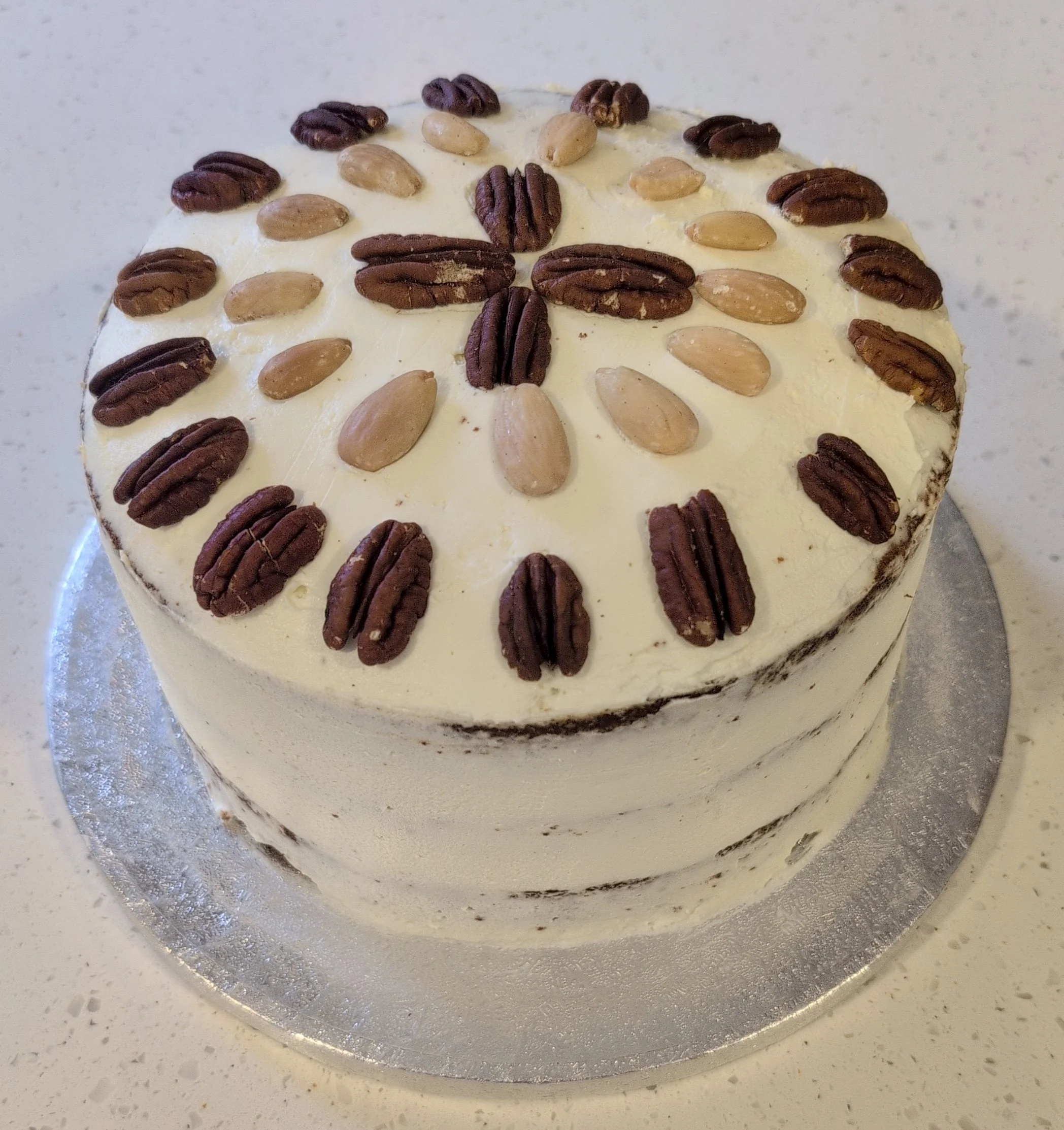A round cake with white frosting decorated with pecans and almonds on top, placed on a silver cake board.
