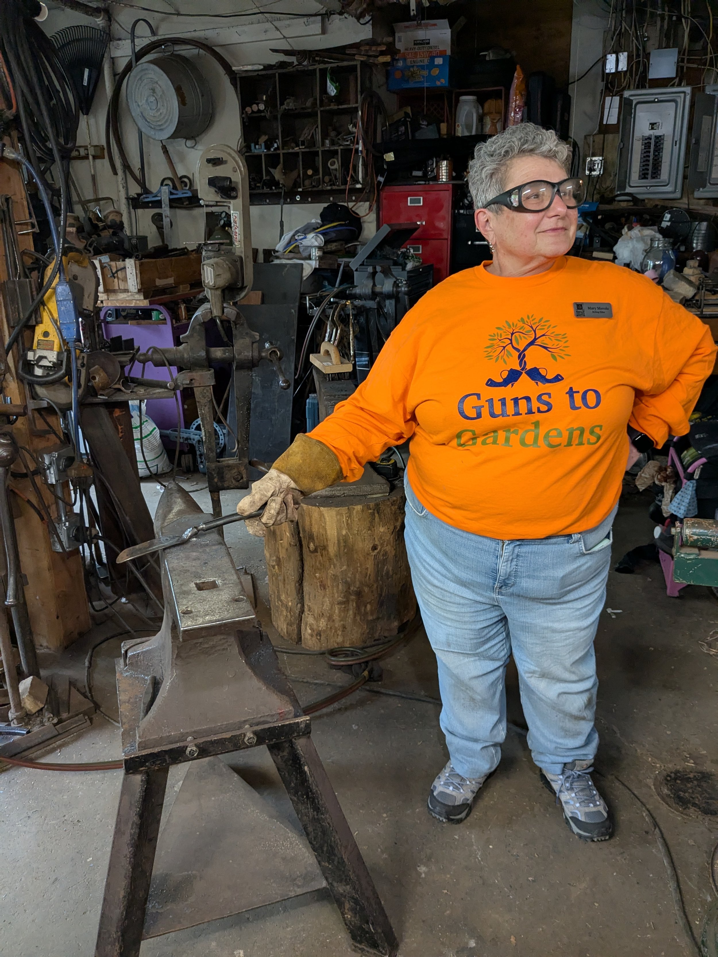 Woman with gray hair and glasses wearing an orange shirt that reads "Guns to Gardens" and jeans, standing in a workshop or garage, holding a metal tool in her right hand, surrounded by various tools and equipment.