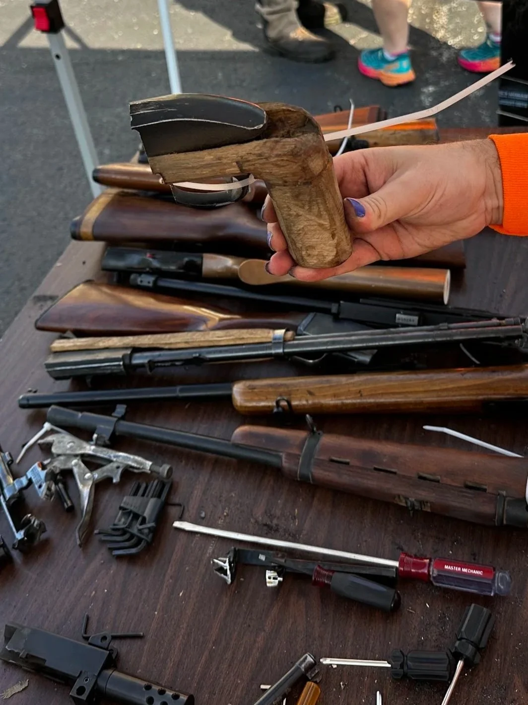 A person in an orange shirt holding a wooden and black rivet gun at an outdoor table displaying disassembled firearms and firearm parts, with other people and vehicles in the background and yellow caution tape nearby.