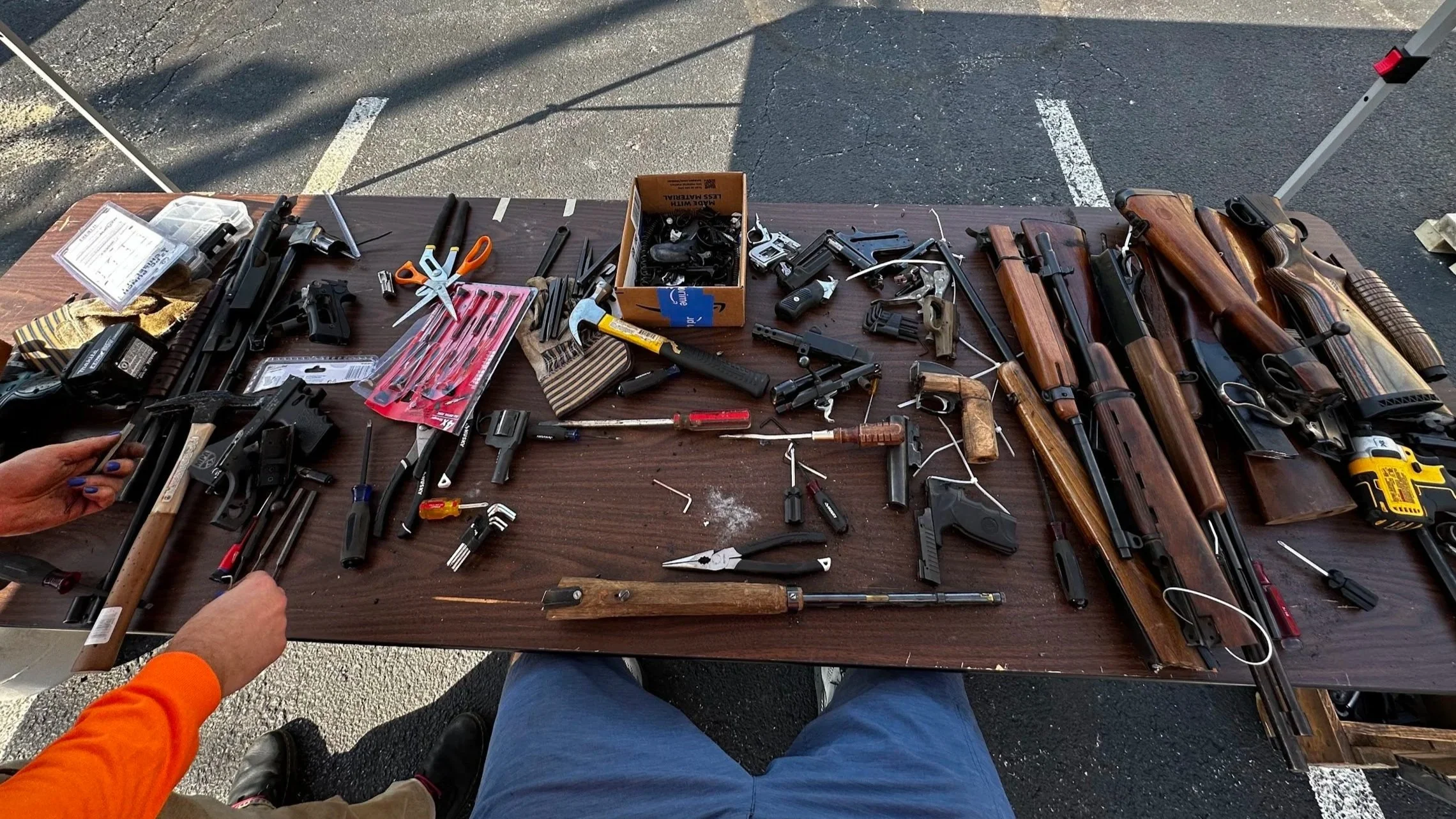 A table with various disassembled firearms, gun parts, tools, and accessories, with two people working on the firearms, in an outdoor setting with yellow caution tape in the background.