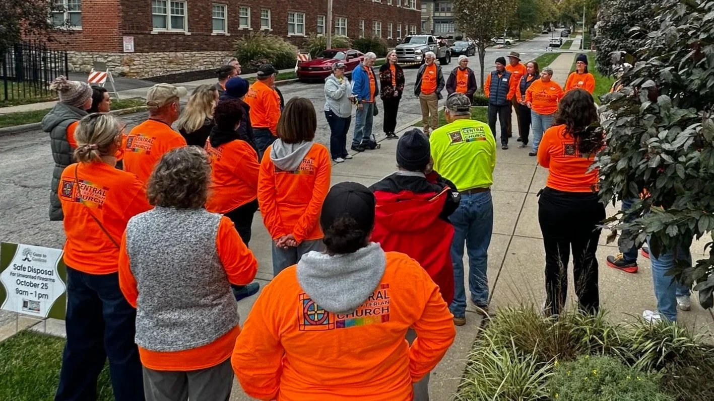 A group of people wearing orange shirts and a few wearing yellow and gray vests are standing on the sidewalk in a circle, participating in a community or volunteer event on a cloudy day. There are residential buildings and trees in the background.