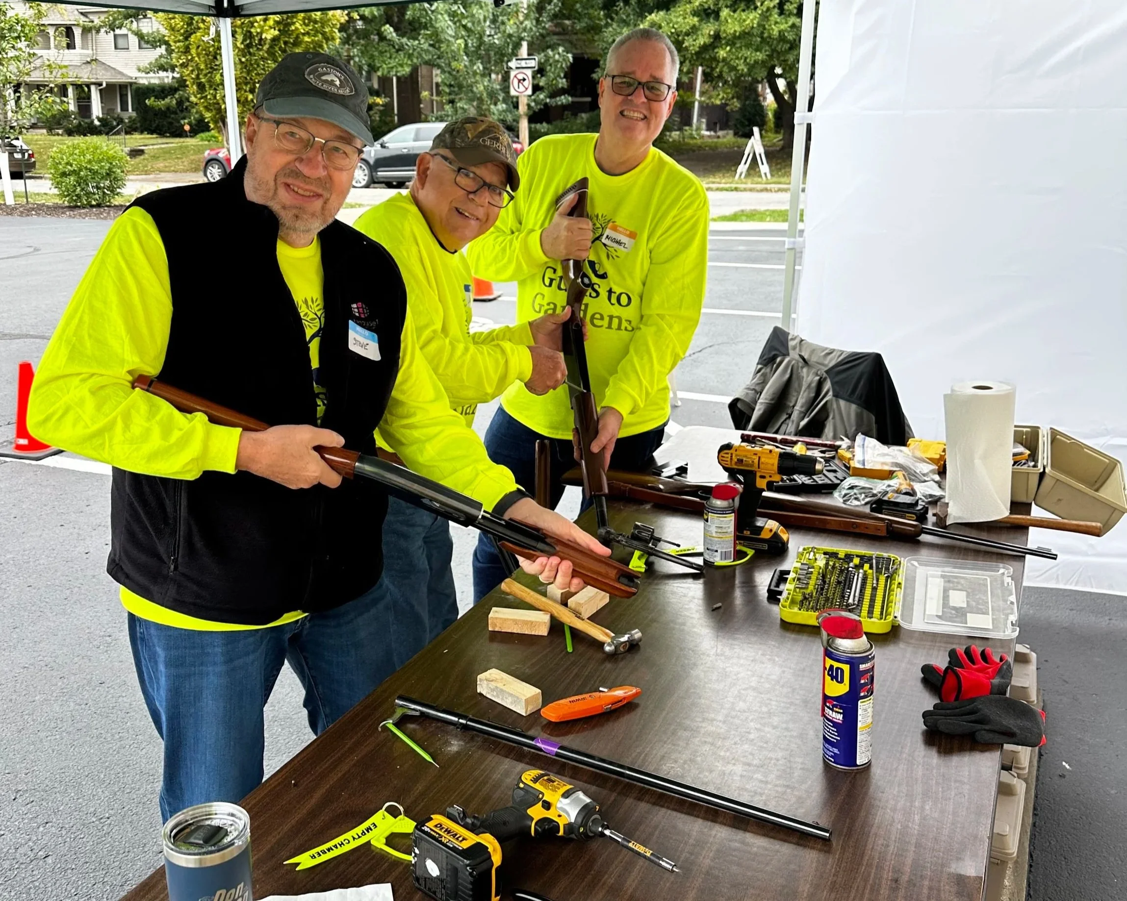 Three men in bright yellow shirts helping to fix rifles under a white tent in a parking lot.