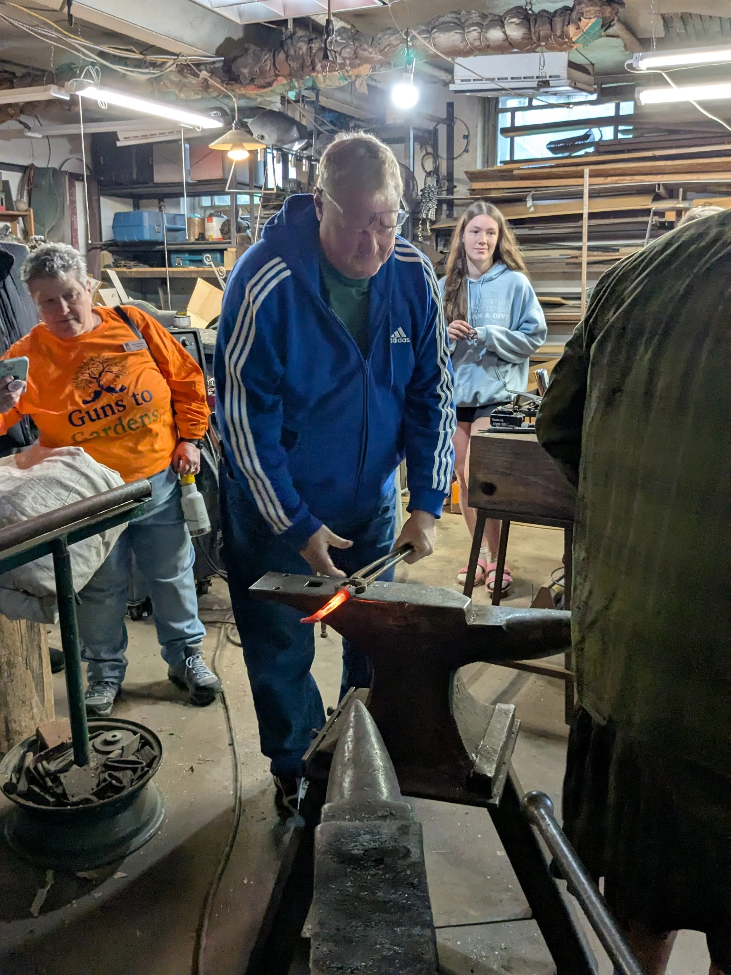 A man wearing a blue Adidas tracksuit is blacksmithing, heating metal on an anvil in a workshop. Inside, there are shelves filled with wood and tools, and a group of women and another man watching him.