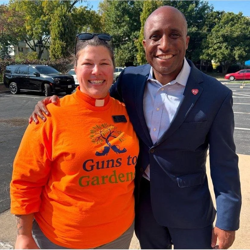 A woman wearing an orange shirt with the slogan 'Guns to Garden' and a lanyard, standing next to a man in a navy blazer and light blue shirt, both smiling outdoors with trees and parked cars in the background.