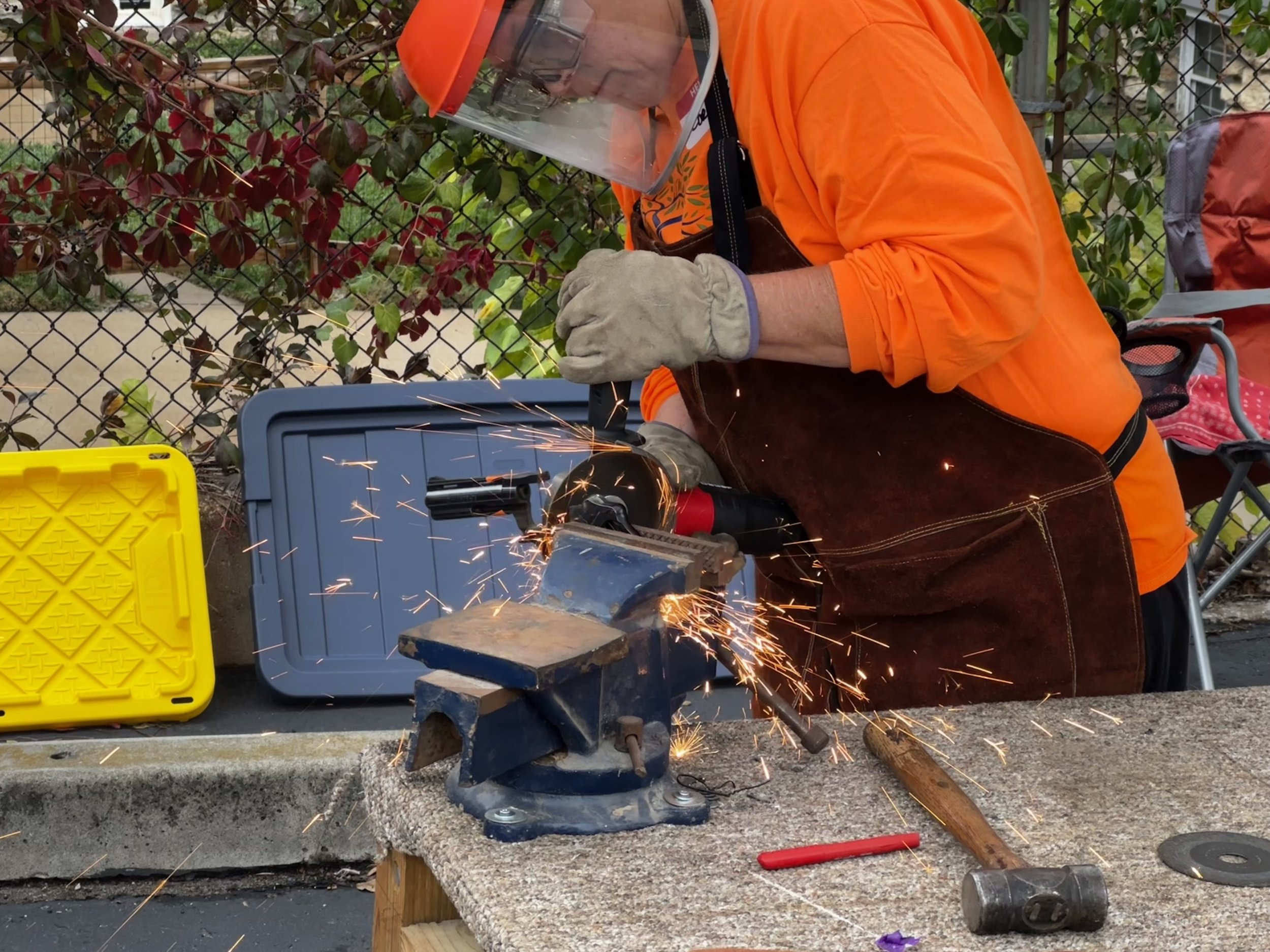 A person wearing an orange shirt, gloves, and a face shield is cutting metal with a grinder, causing sparks. There are tool cases and a hammer on the work surface, which is a wooden plank covered with a textured material, with a fence and plants in t