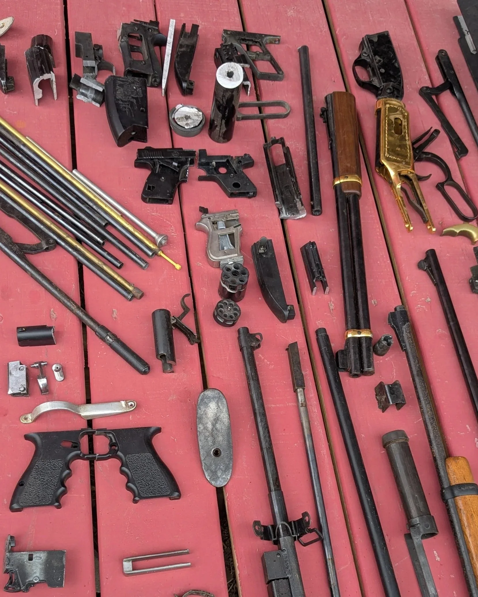 Disassembled firearms parts spread on a red wooden surface, including gun frames, barrels, slides, cylinders, magazines, and other firearm components.
