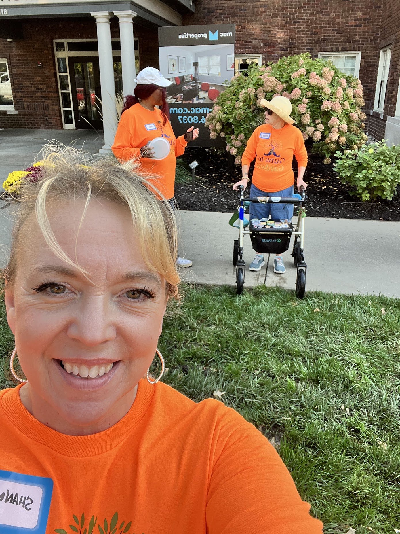 Three women outdoors wearing orange shirts, two women with sun hats, one with a white cap, one woman with glasses, standing in front of a brick building with a large bush. The woman in the foreground is smiling for a selfie, while the other two women