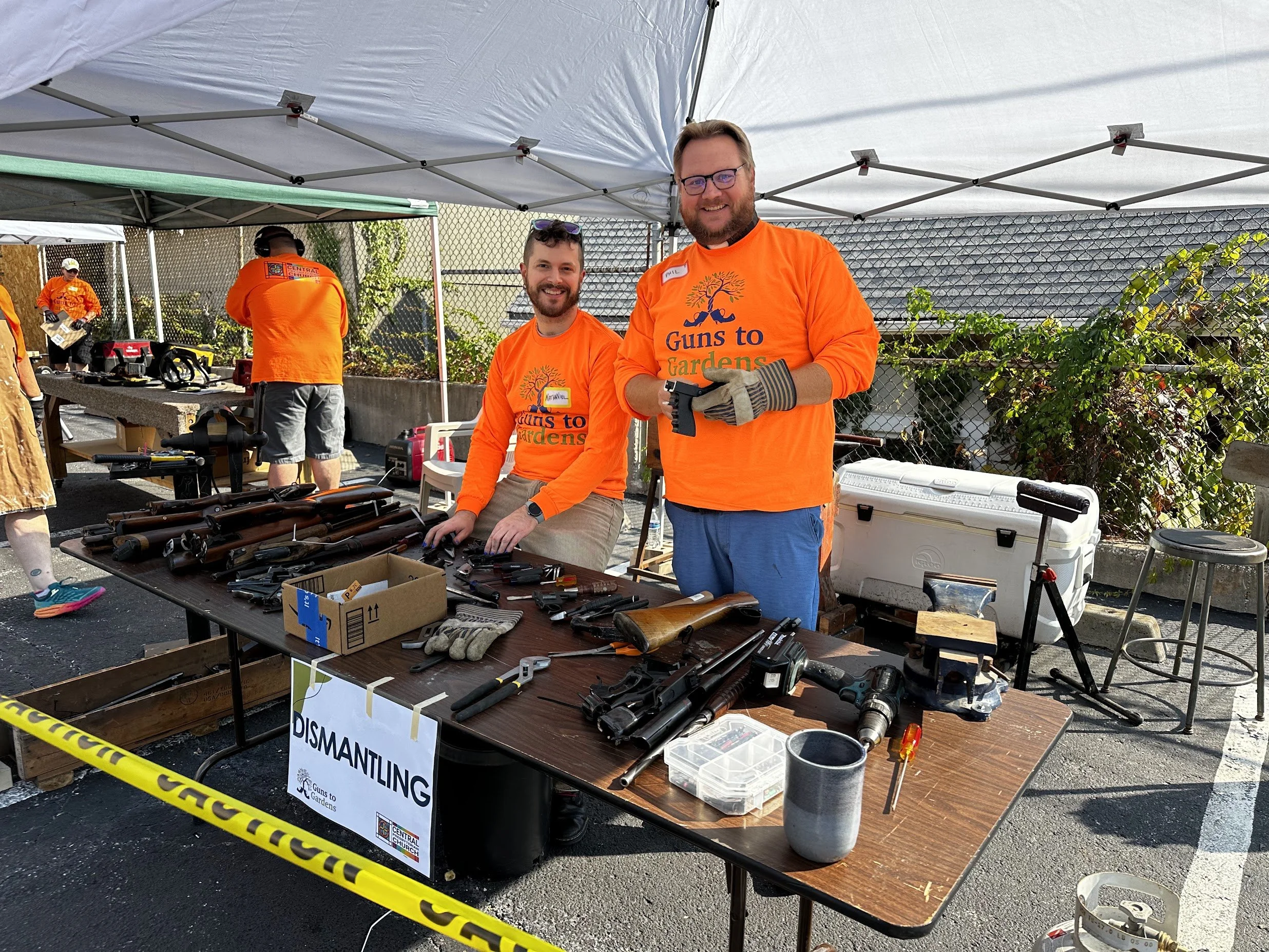 Two smiling men in orange shirts with 'Guns to Gardens' logo, dismantling rifles at an outdoor event table with various tools and disassembled guns, under a tent, with other people in similar shirts in the background.
