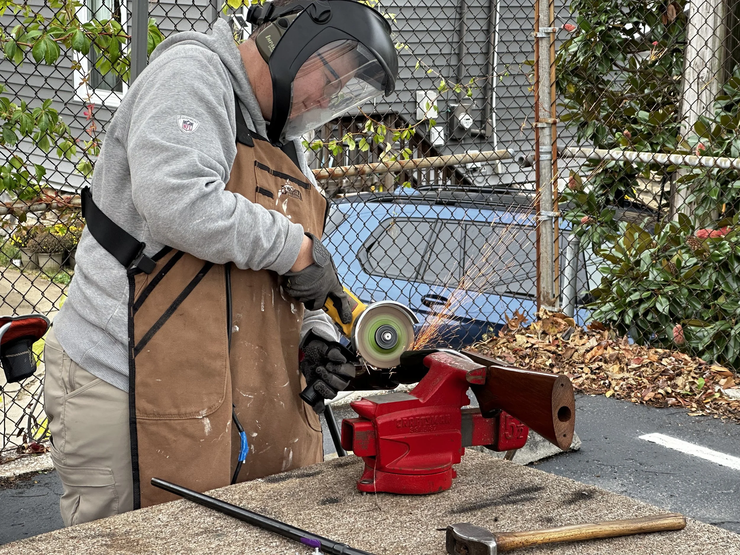 A man wearing a helmet, gray sweatshirt, apron, and gloves is using an angle grinder on a piece of wood secured in a red vise on a workbench outside. Sparks are flying from the grinding action.