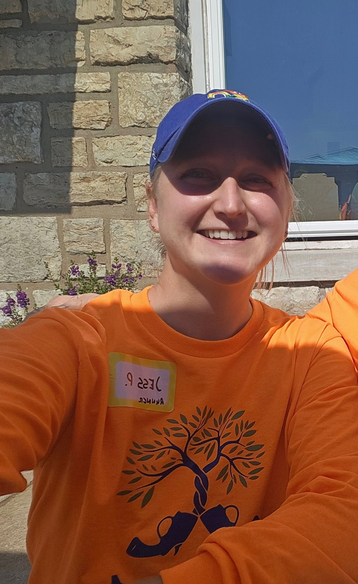 A woman smiling outside, wearing an orange shirt with a tree graphic and a blue cap, with a stone wall and window in the background.