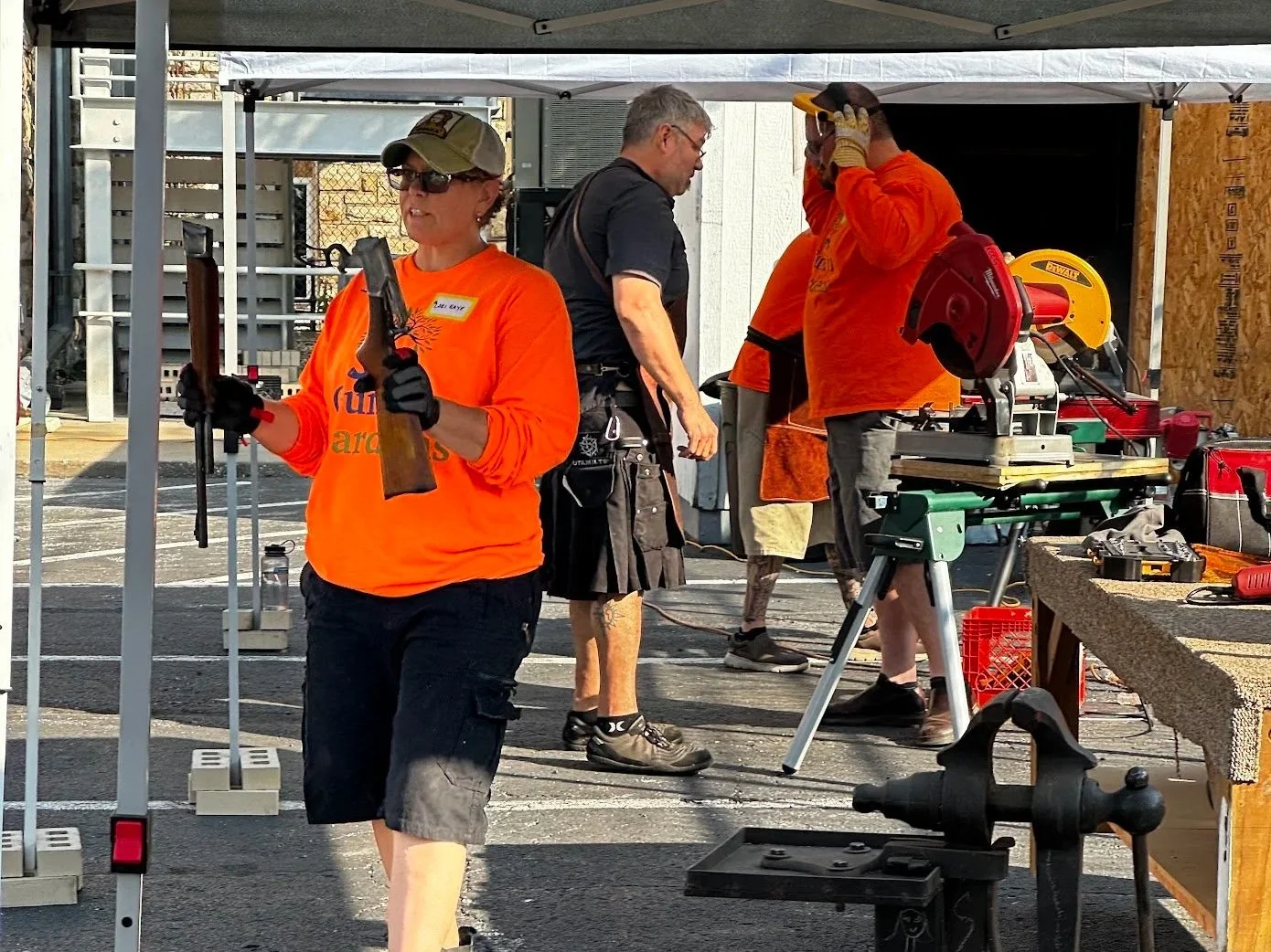 Group of people working outdoors with tools and equipment, some wearing orange shirts and safety gear, setting up for a construction or event project.