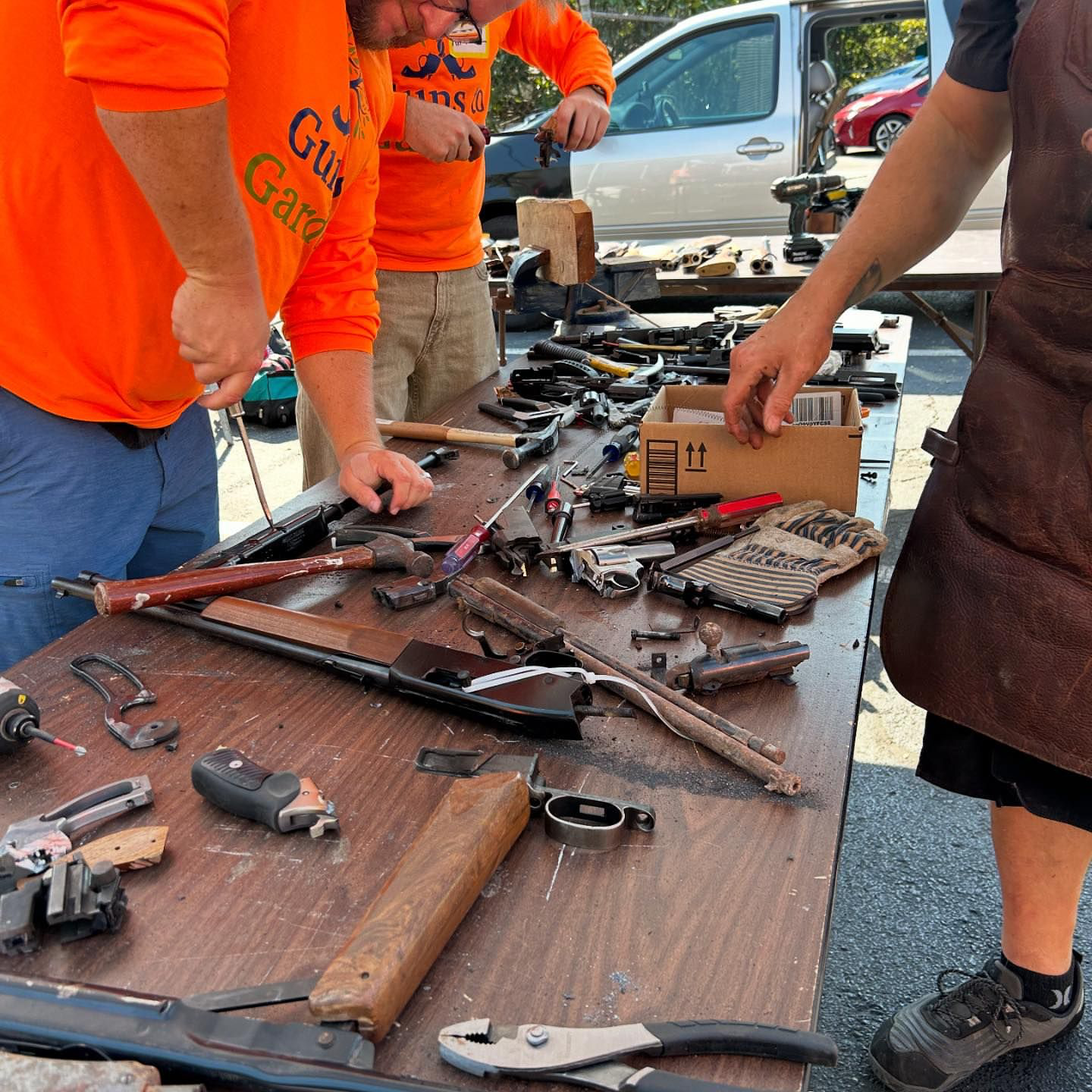 People working on guns at a table filled with various handguns, ammunition, and tools in an outdoor setting.