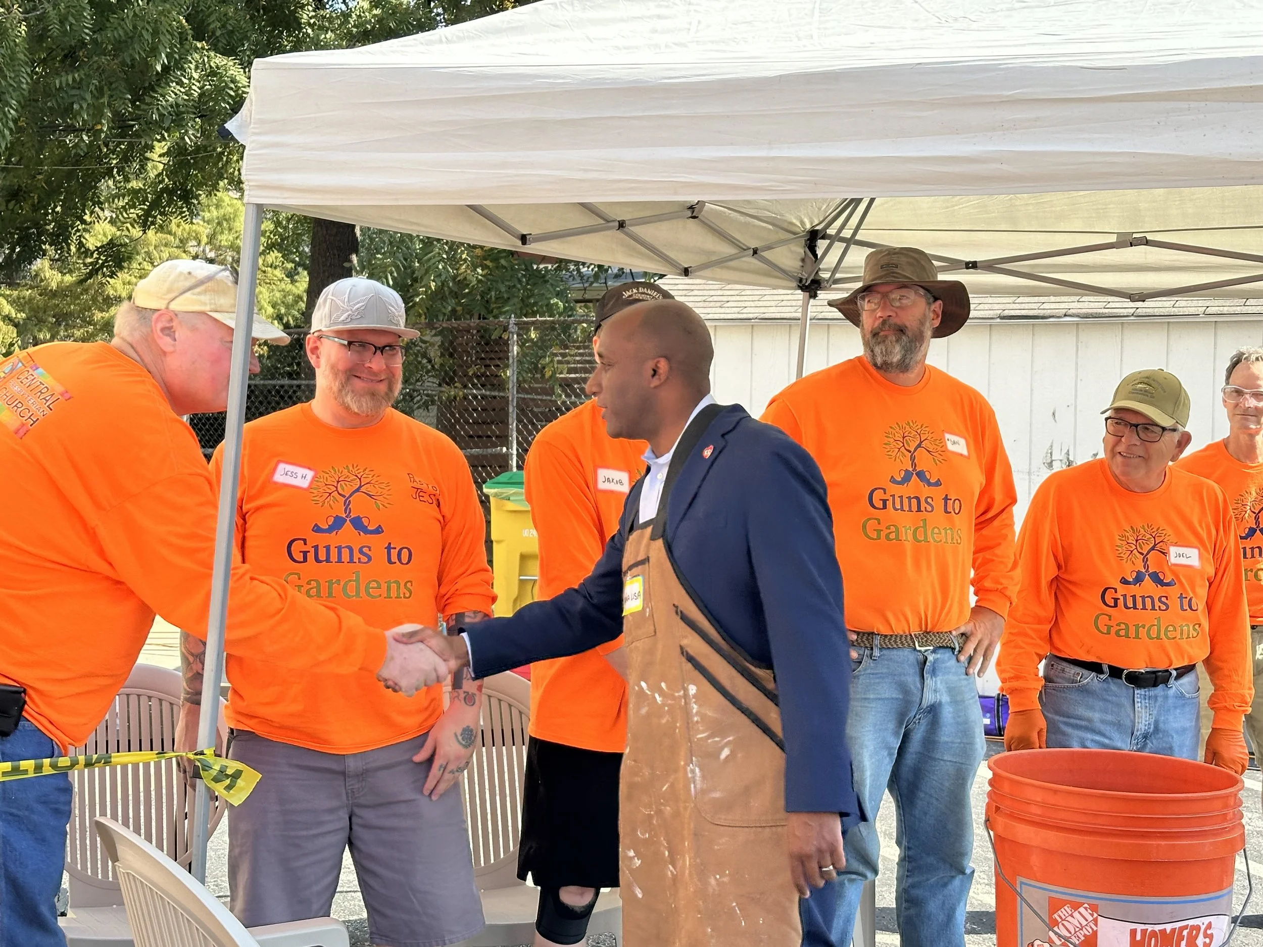 A man in a navy blazer shaking hands with another man wearing an apron, socializing under a white canopy tent with five other men in orange "Guns to Gardens" shirts, outdoors on a sunny day.
