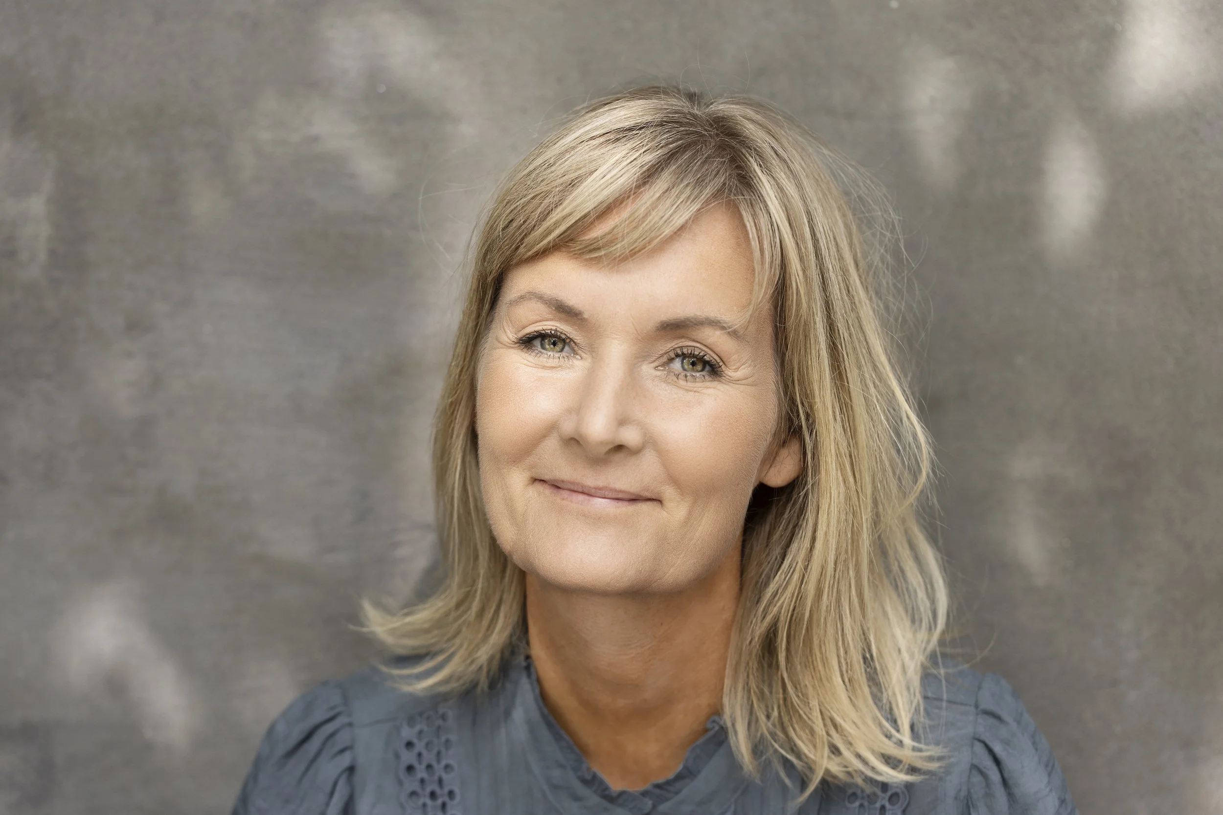 Et portræt af en smilende kvinde med blondt hår og grønne øjne, iført en blå bluse, foran en glat gray baggrund.