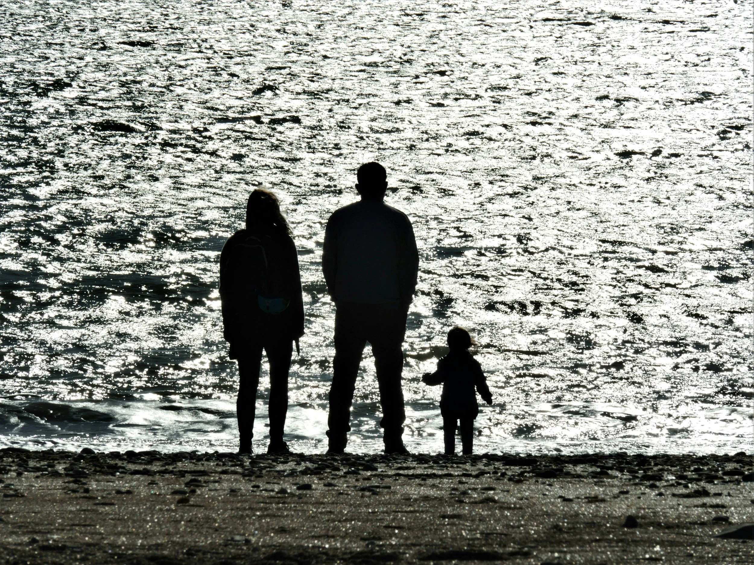 Silhuetter af en voksen, en teenager og to børn, der går mod havet ved solnedgang, hvor havet reflekterer sollyset.