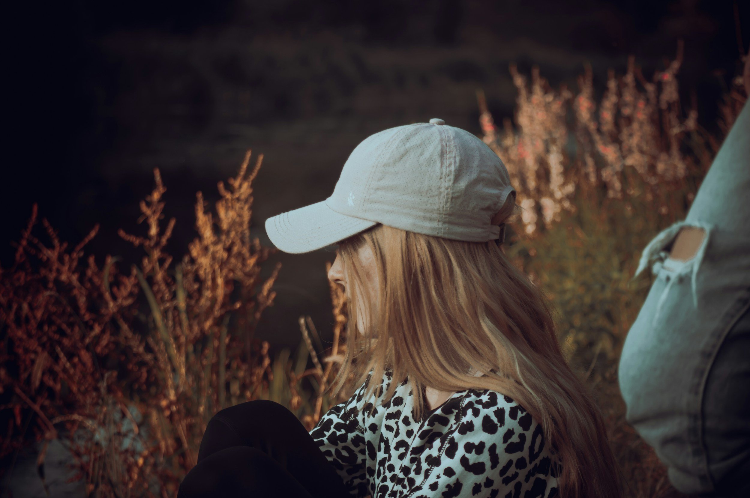 En person med langt blondt hår, der sidder ved siden af en person i en grå jakke. Personen med blondt hår har på en lysegrå Cap, og de er udendørs med buskads, der har brune blade, bag dem i skumringen.
