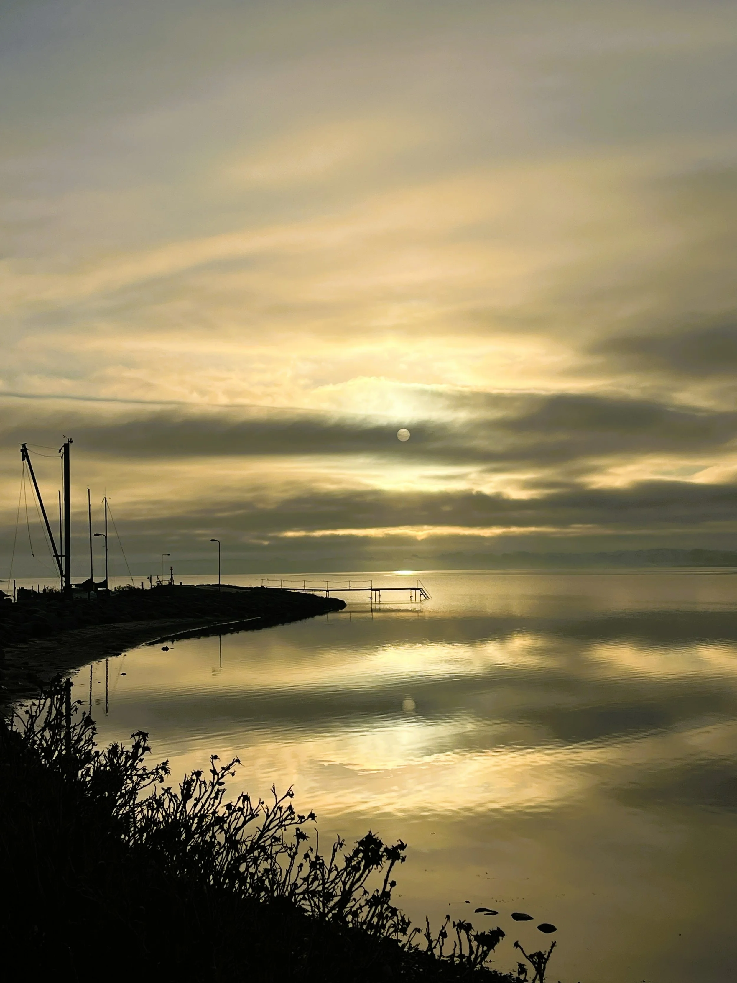 Solnedgang over en rolig havn med reflekterende vand, få klippekæder og nogle bådstænger i forgrunden.