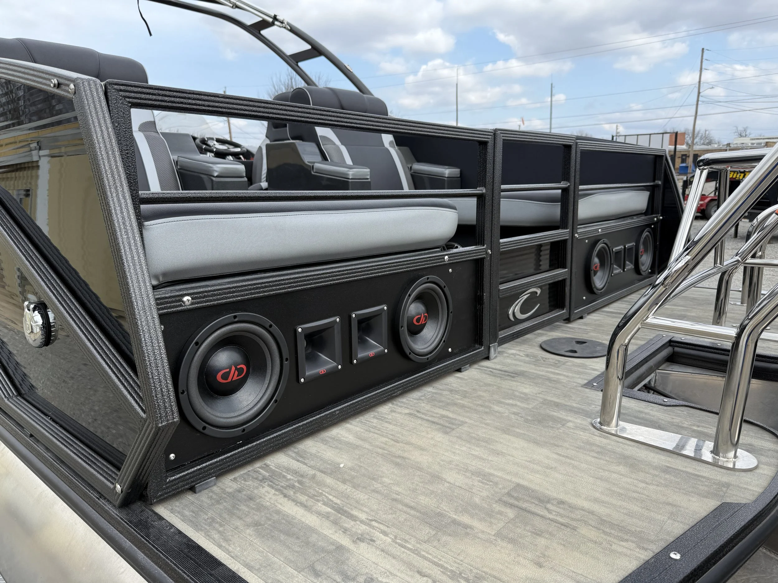 Close-up of a boat's deck with a custom audio system, including multiple speakers and subwoofers, surrounded by seating and metal rails, with a cloudy sky and power lines in the background.