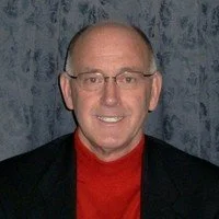 A smiling man with glasses, wearing a red shirt and black blazer, in front of a textured blue background.