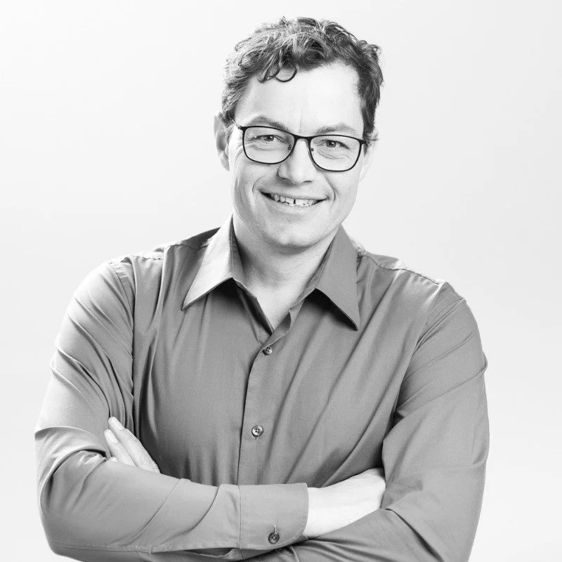 Black and white photo of a smiling man with curly hair, wearing glasses and a button-up shirt, crossing his arms.