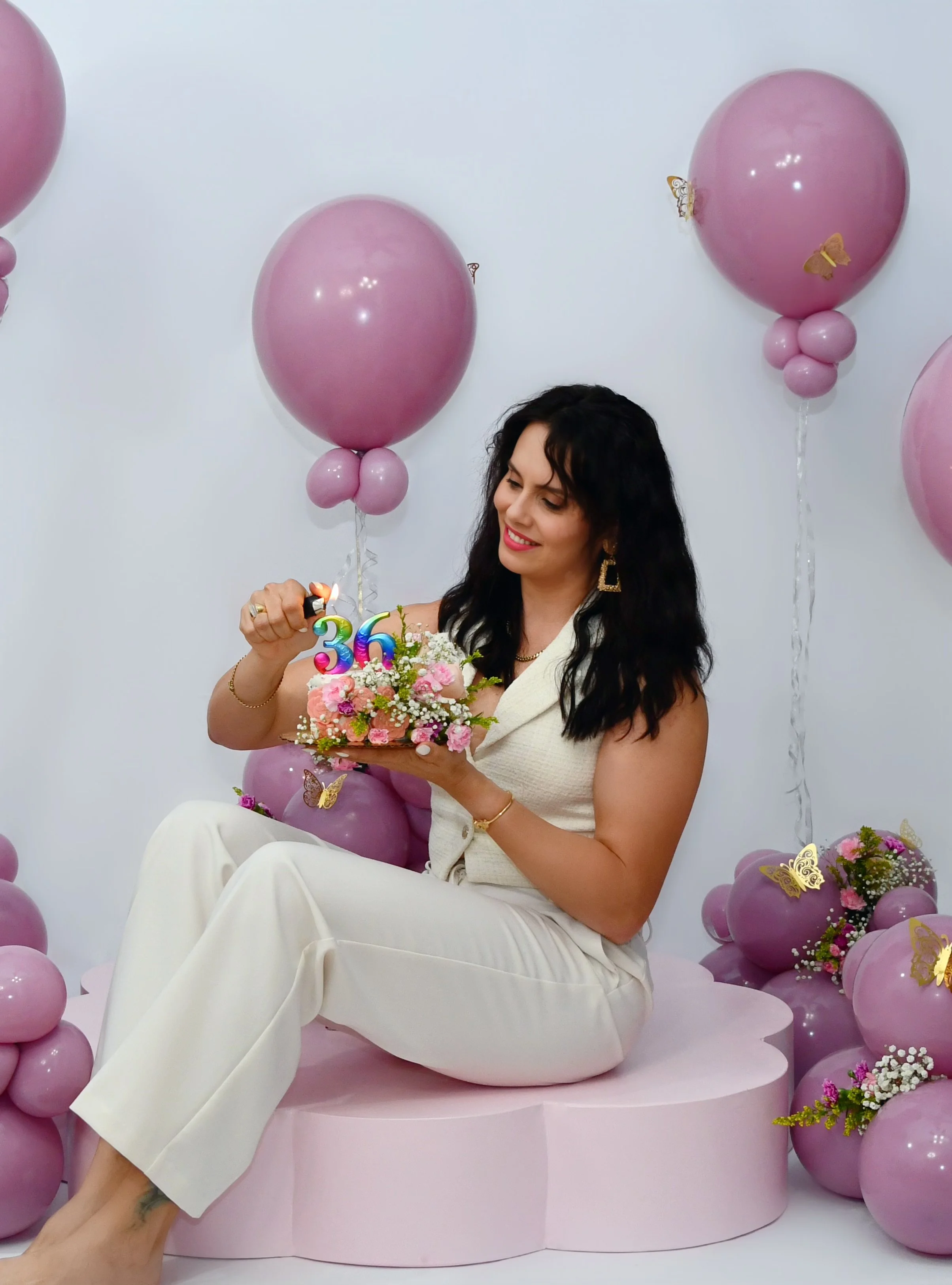 A woman sitting on a pink circular platform, smiling while holding a bouquet of flowers and a birthday cake with colorful candles that spell out 36. She is surrounded by pink balloons and butterfly decorations.