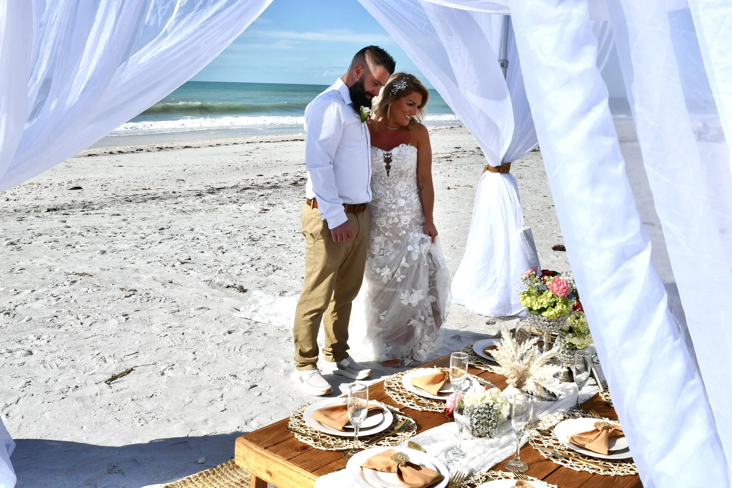 A couple getting married on the beach, standing under a white canopy near a table set with plates, glasses, and flowers.