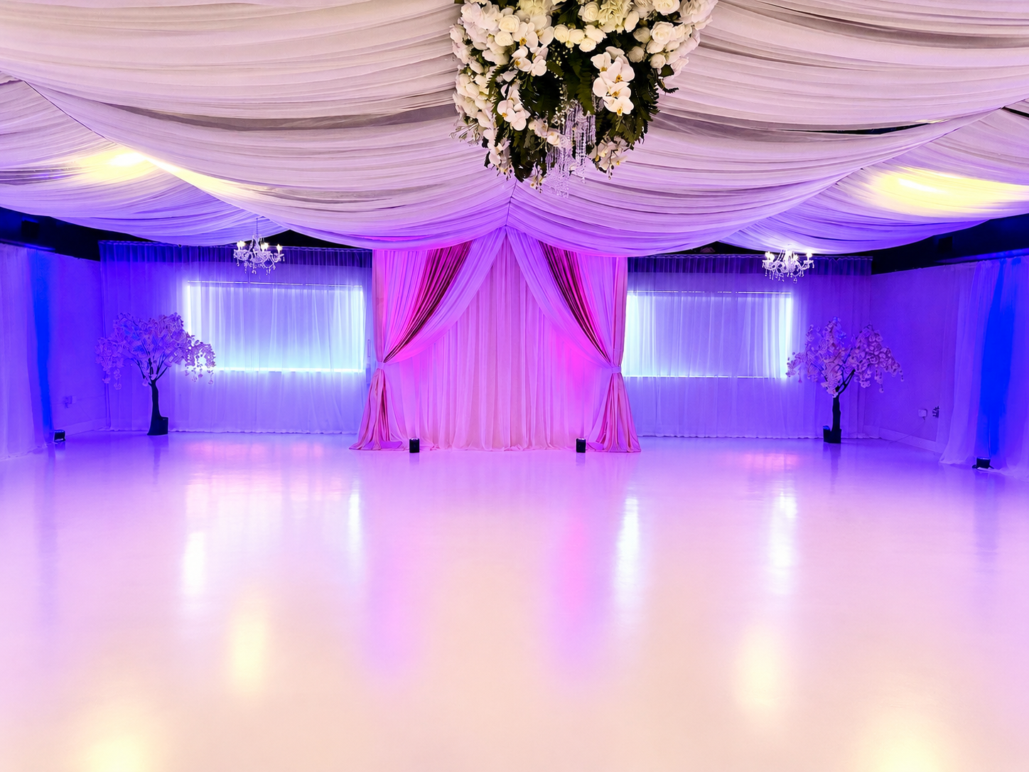 Elegant event space decorated with flowing pink and purple drapes, floral arrangements, and chandeliers, illuminated with soft pink, purple, and blue lighting.