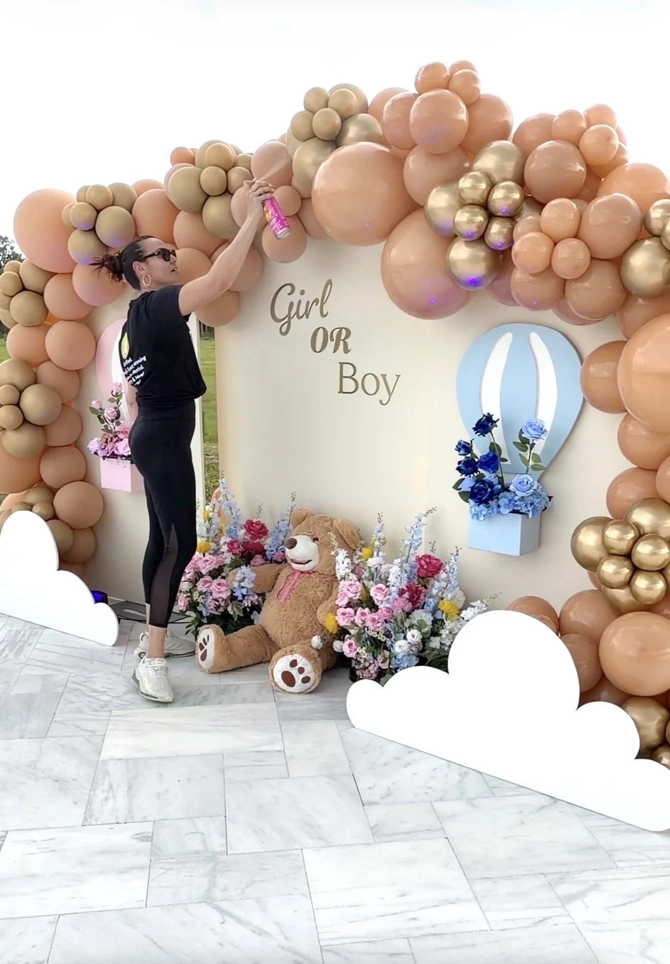 A woman in black athletic clothing and sunglasses is standing next to a decorative backdrop made of pink, gold, and peach balloons, with the words "Girl OR Boy" in the center. The woman is holding a can of spray while looking up. At the base of the backdrop, there is a large plush teddy bear surrounded by pink and blue flowers, with white cloud cutouts in the foreground.