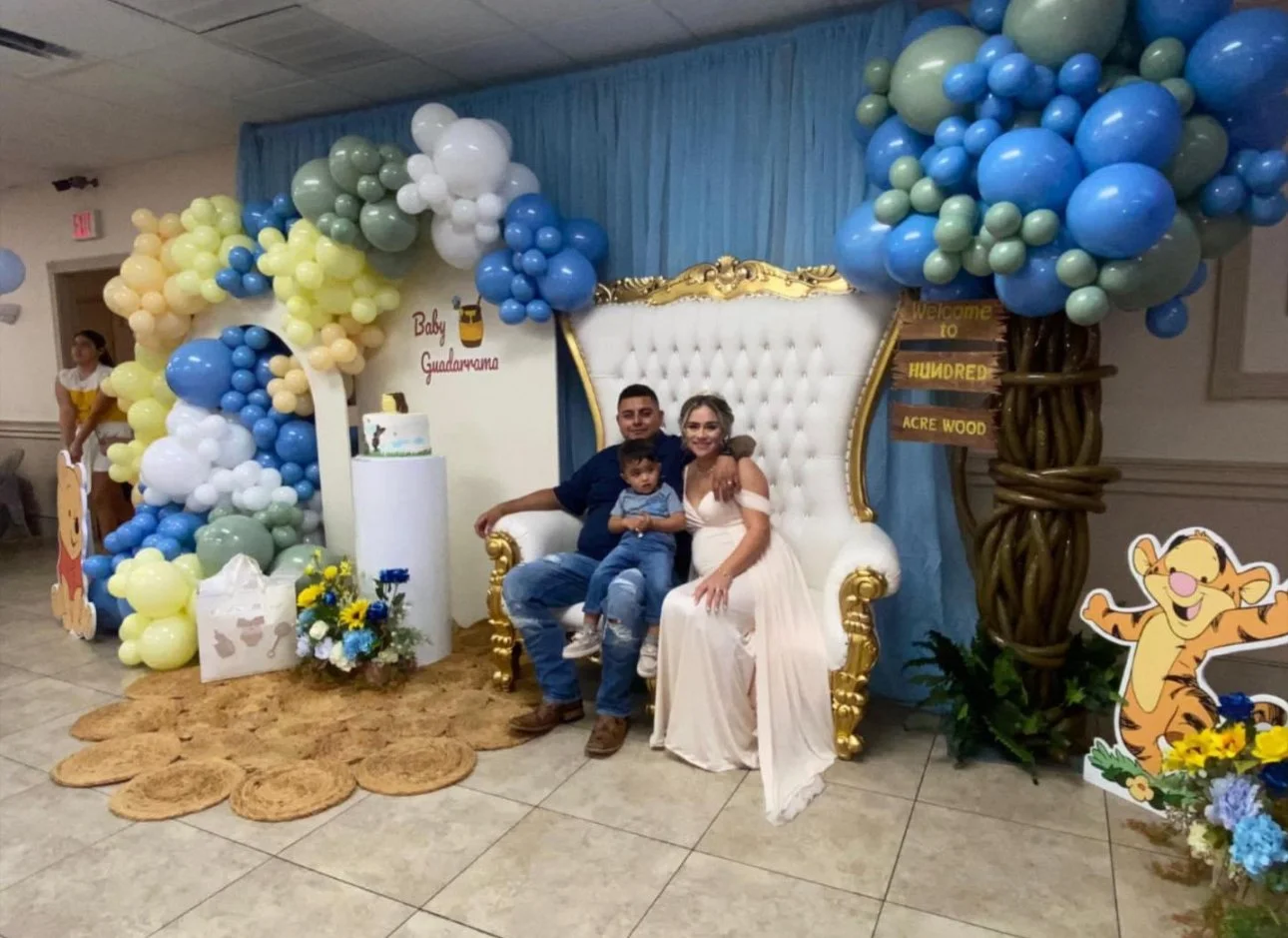 Family sitting on a white and gold throne with blue and yellow balloon decorations, a cake, and tiger-themed decorations for a baby shower celebrating Baby Guadarrama.