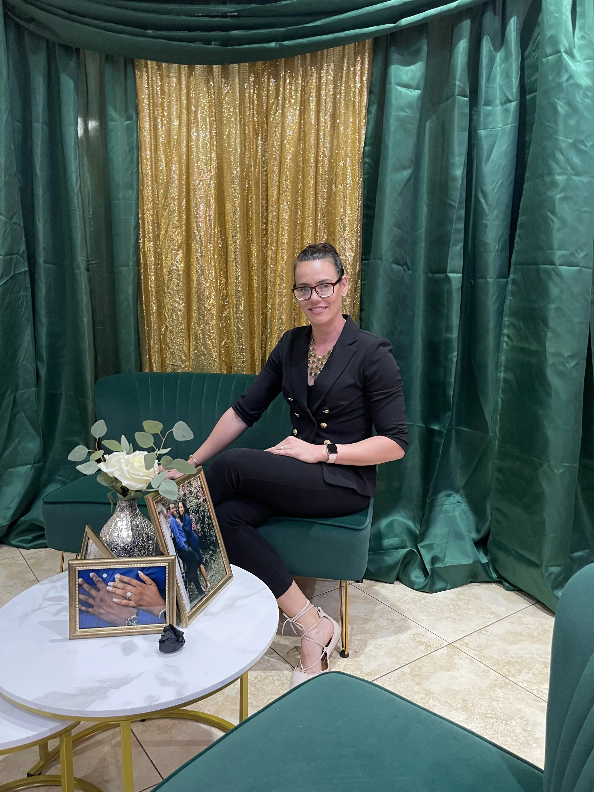 A woman in a black blazer and pants, with glasses, sitting on a green chair in front of a backdrop of green curtains and a gold curtain. A white table with framed photographs and a floral arrangement is in the foreground.