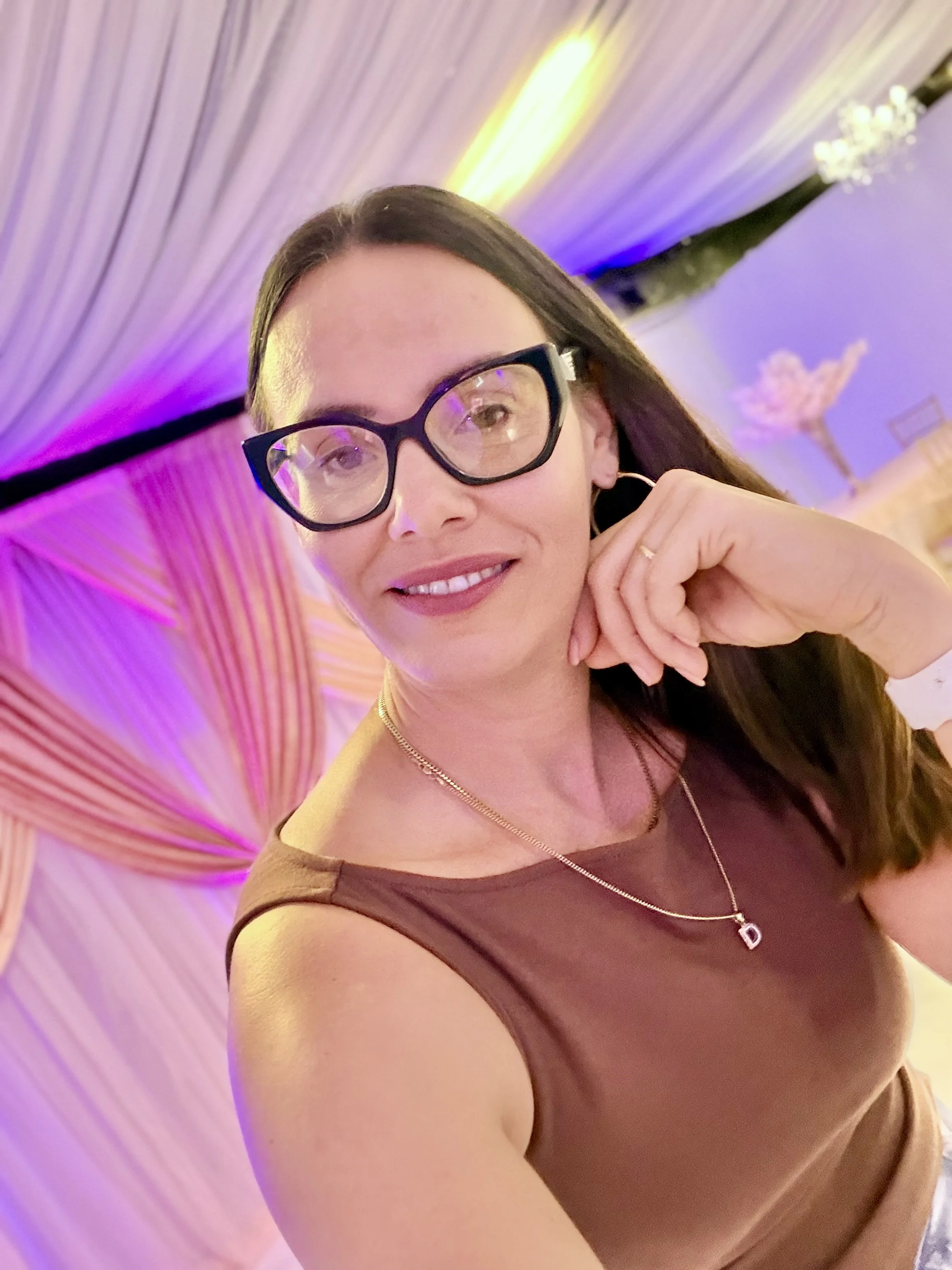 A woman with brown hair and large black glasses taking a selfie at a decorated event with colorful, illuminated drapes and floral decorations in the background.