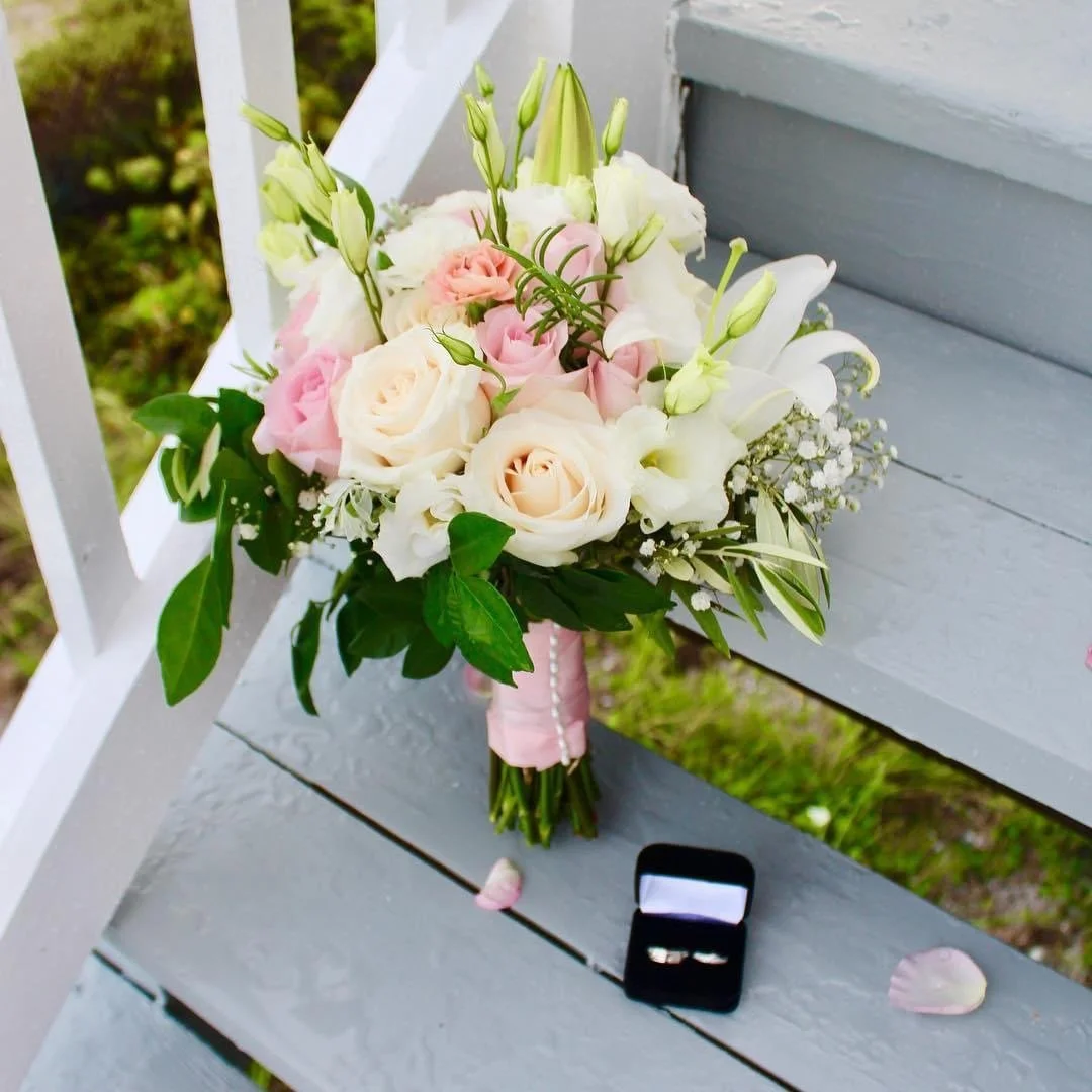 A bridal bouquet of white and light pink roses, white lilies, and greenery, with a pink ribbon around the stems, resting on a gray wooden bench next to a black ring box with rings inside and scattered pink rose petals.