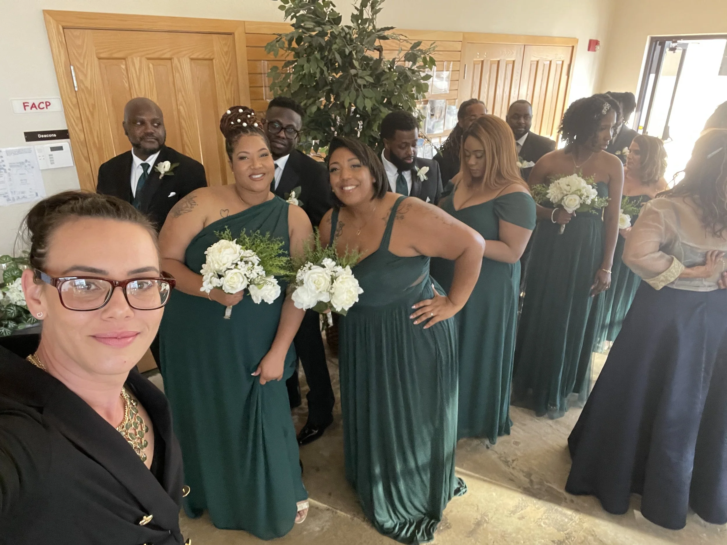 A group of wedding party members, including bridesmaids in green dresses holding white bouquets and groomsmen dressed in black suits with teal ties, standing indoors near wooden walls and a door, with a woman taking a selfie in the foreground.