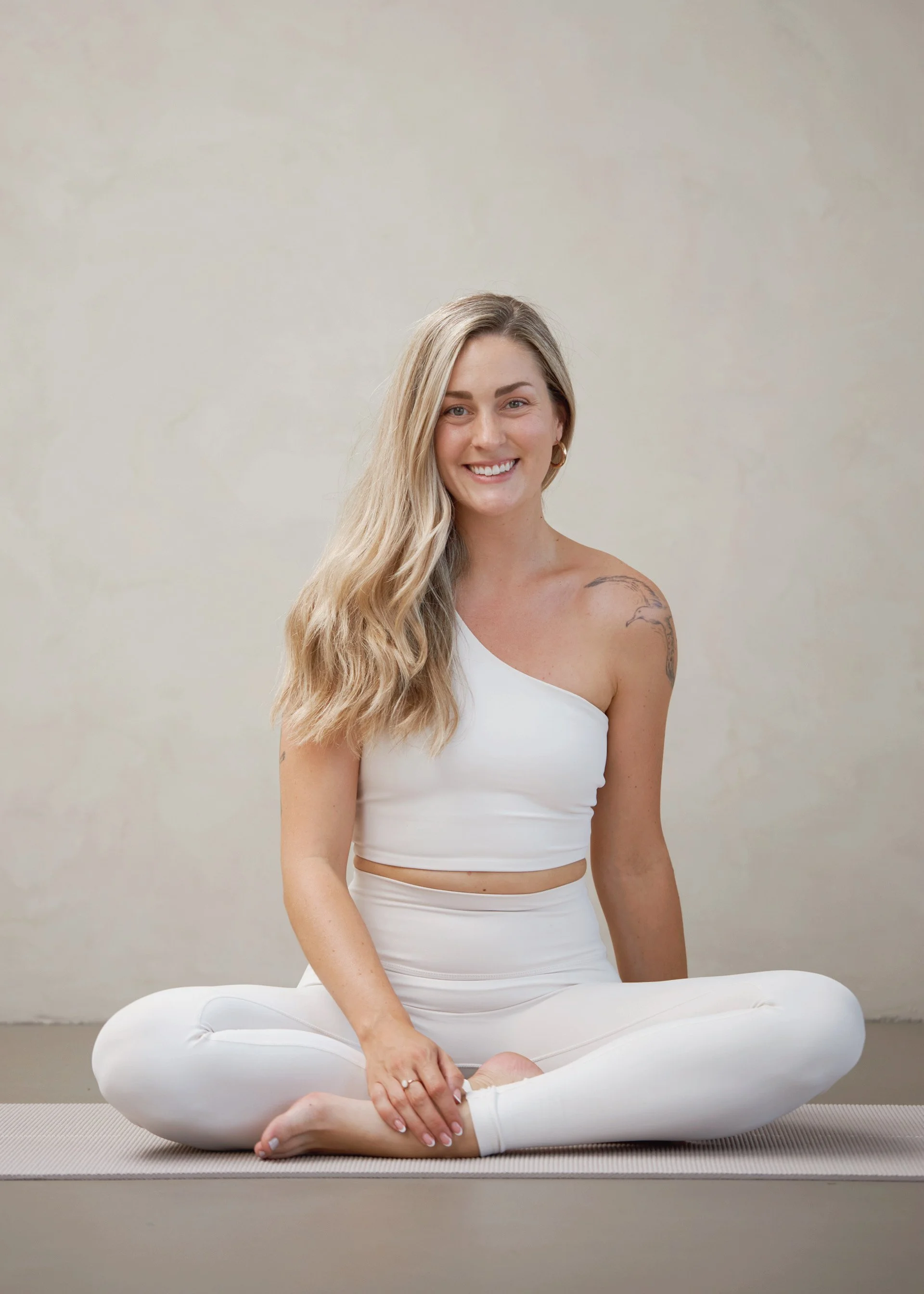 A woman with long blonde hair and a tattoo on her right shoulder, sitting cross-legged on a yoga mat, smiling at the camera, wearing a white one-shoulder top and white leggings.