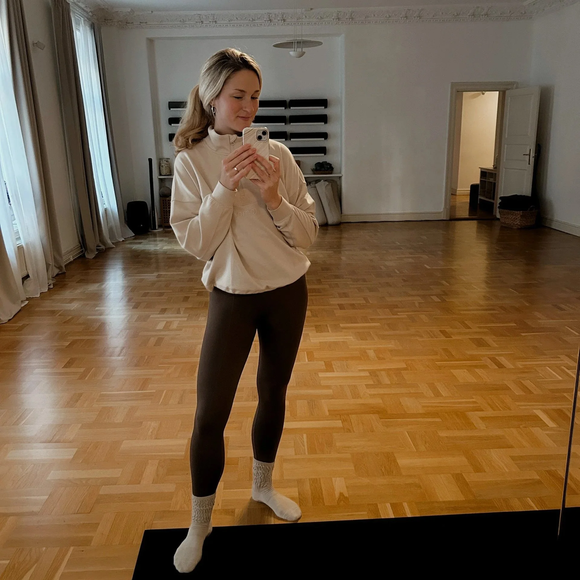 En ung kvinna tar en selfiespegelbild i ett tomt rum med trägolv. Hon bär en beige tröja, svarta leggings och vita sockor.
