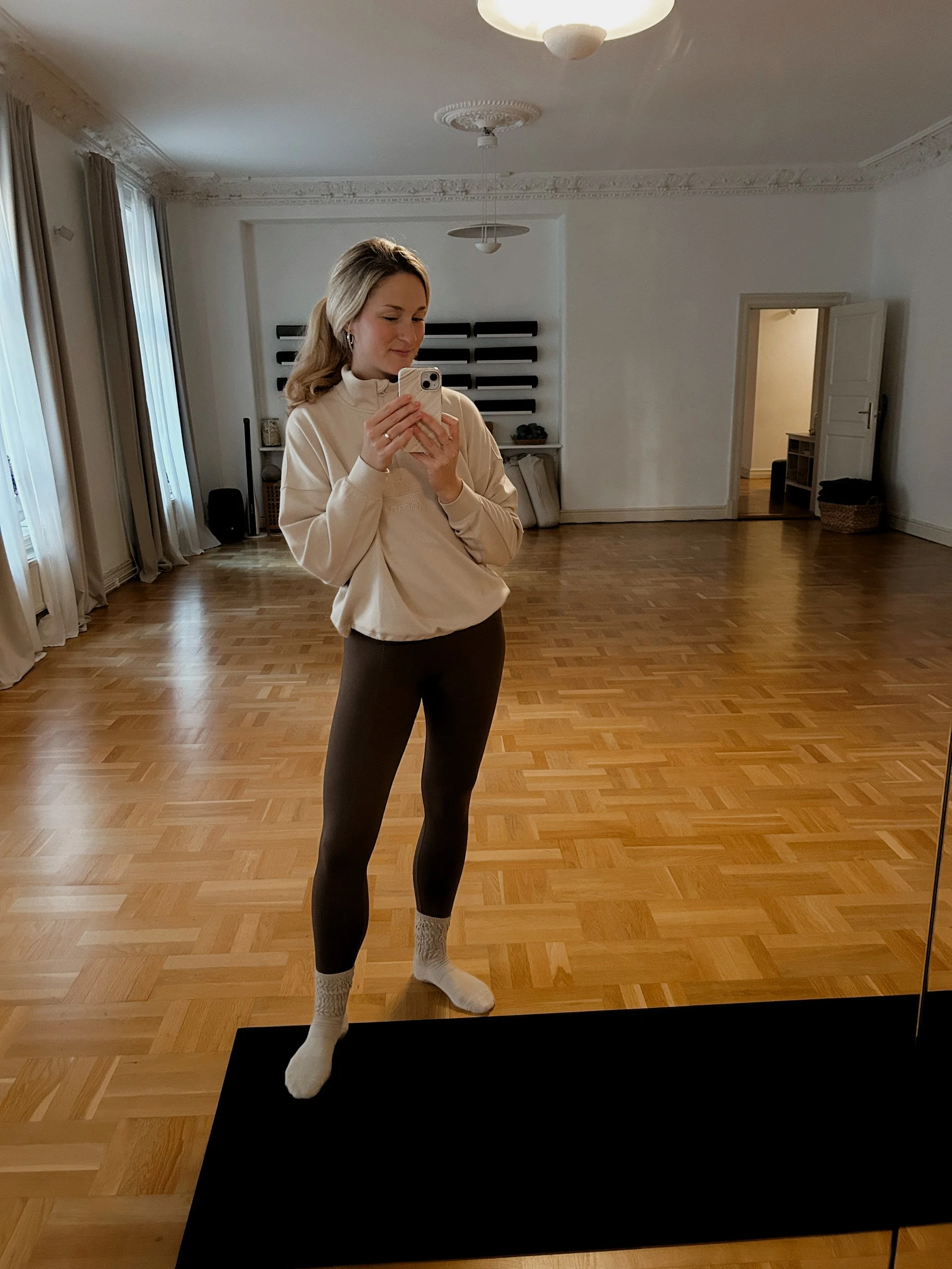 En ung kvinna tar en selfie i ett tomt rum med parkettgolv, vita väggar och stora fönster med vita gardiner. Hon har blont, lockigt hår och bär en beige sweatshirt, svarta tights och vita sockor.