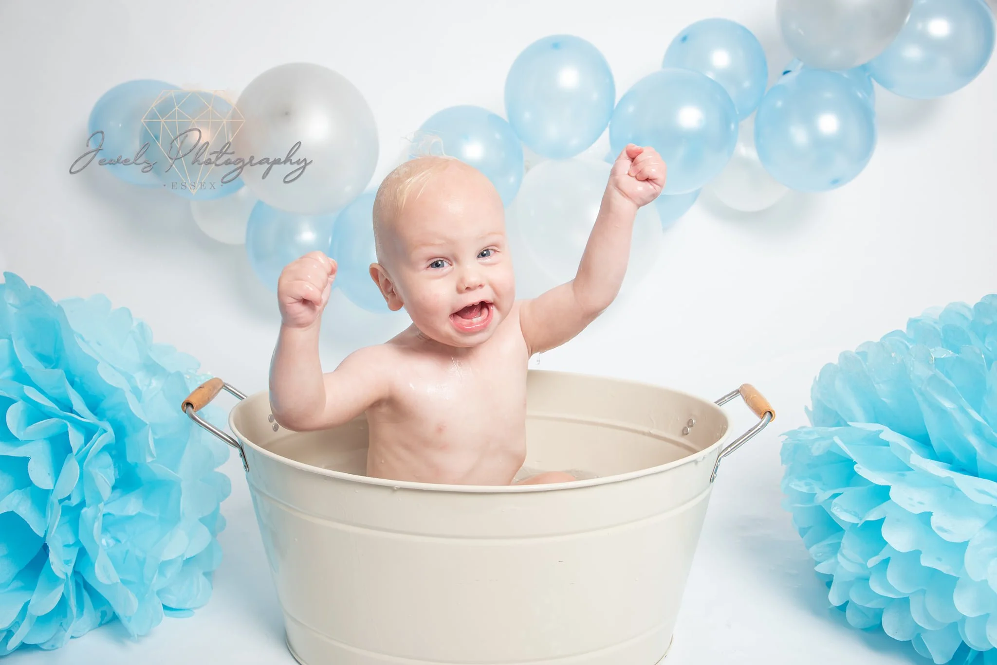 A smiling baby with wet hair sitting in a white bucket, raising both fists in celebration, surrounded by blue and white balloons and blue tissue paper pom-poms.