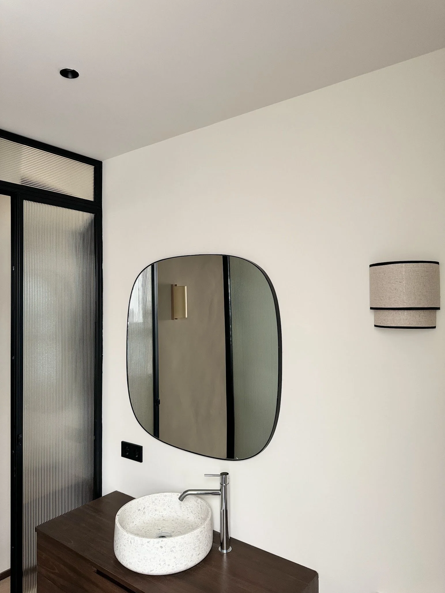 Modern bathroom with a white stone vessel sink, a round black-framed mirror, and a beige wall-mounted light fixture.