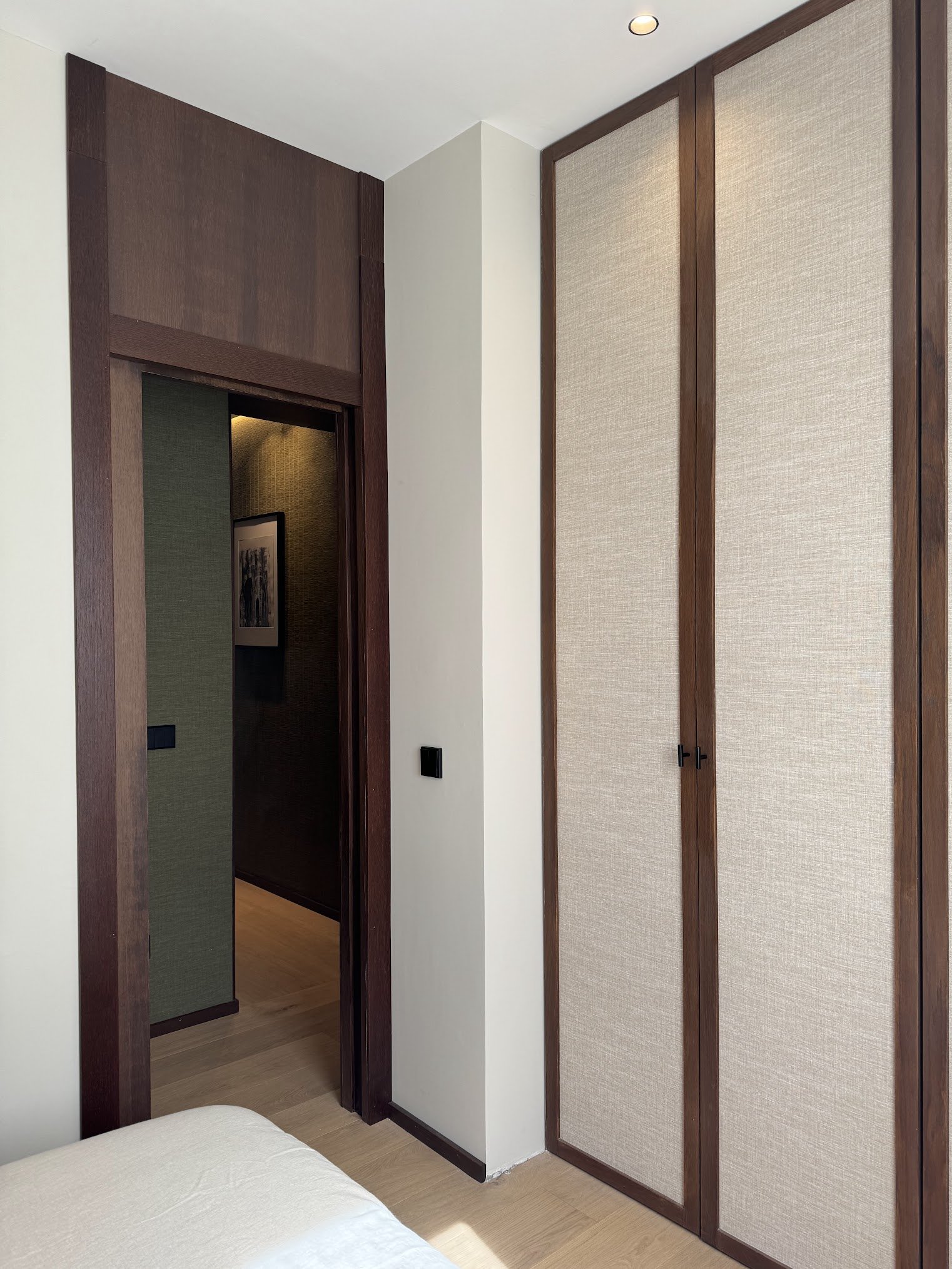 Interior of a bedroom showing a white bed, a doorway with wooden trim leading to a hallway, and built-in wardrobes with beige fabric doors.
