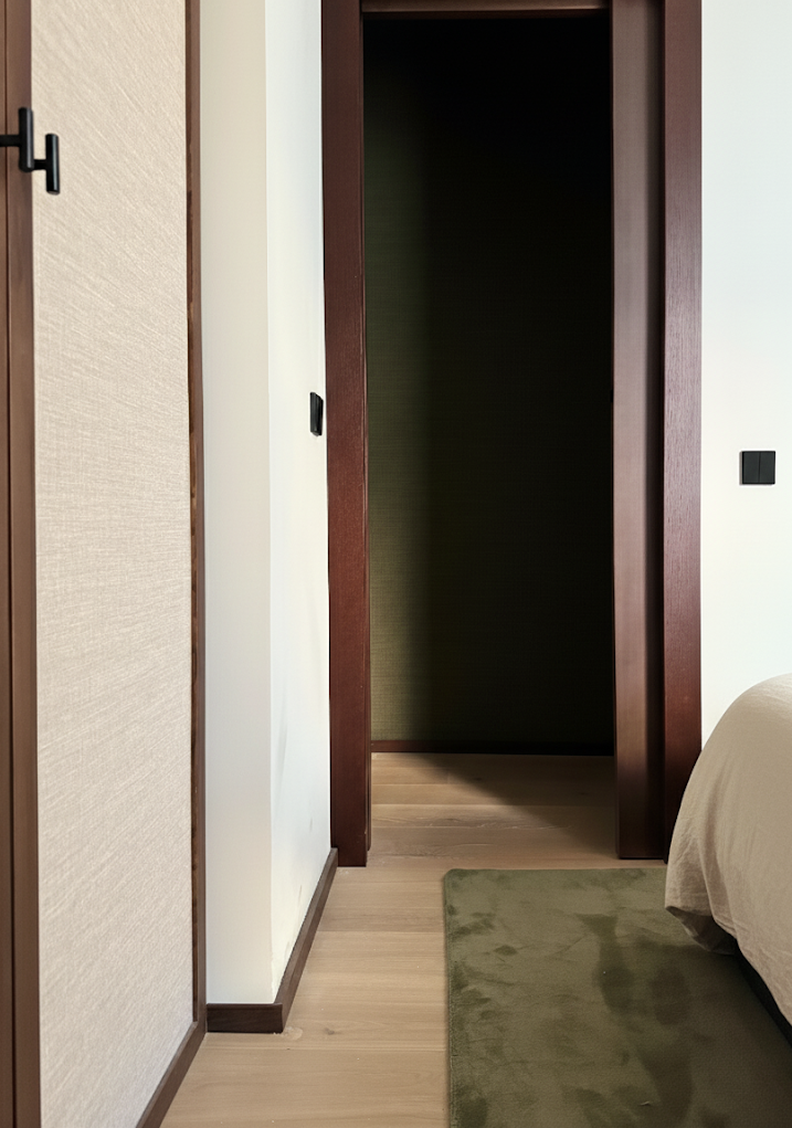 A bedroom with a wooden floor, a green rug, and a bed partially visible on the right. There is a closet with a sliding door and a doorway to another room or hallway in the background.