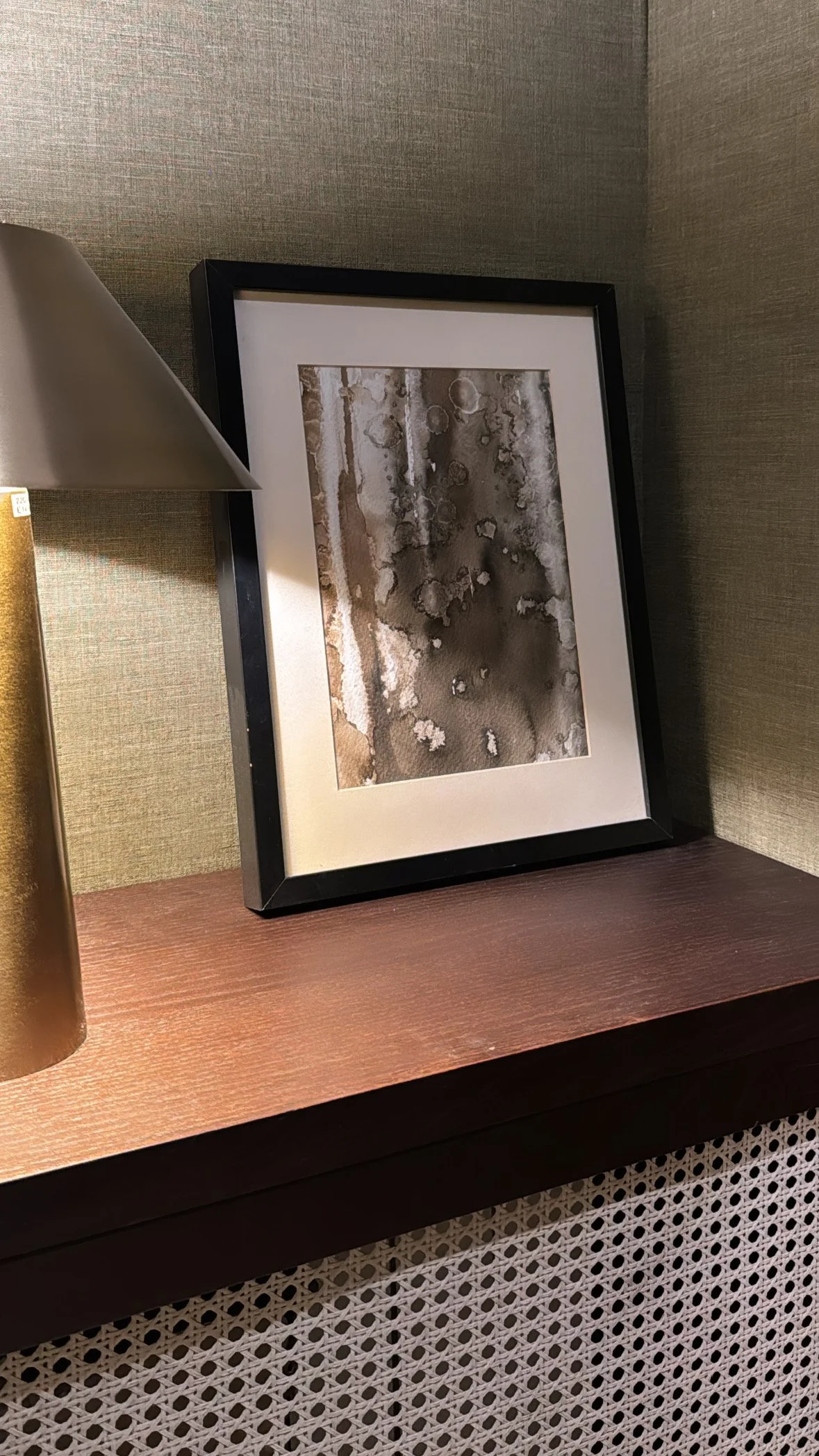 Framed abstract black and white watercolor painting on a wooden shelf, partially lit by a nearby lamp.