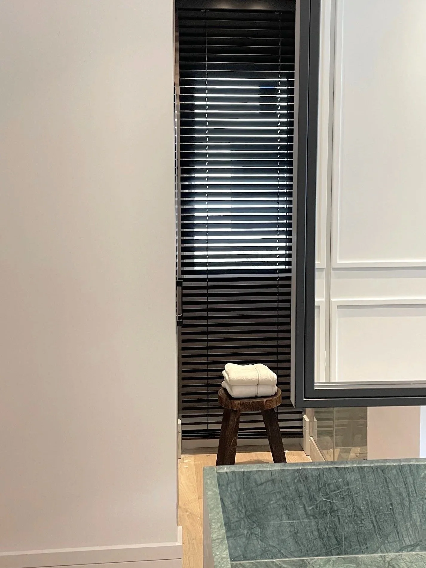 A small wooden stool with folded white towels on top, positioned beside a kitchen island with a greenish countertop, near a wall with black window blinds partially open.