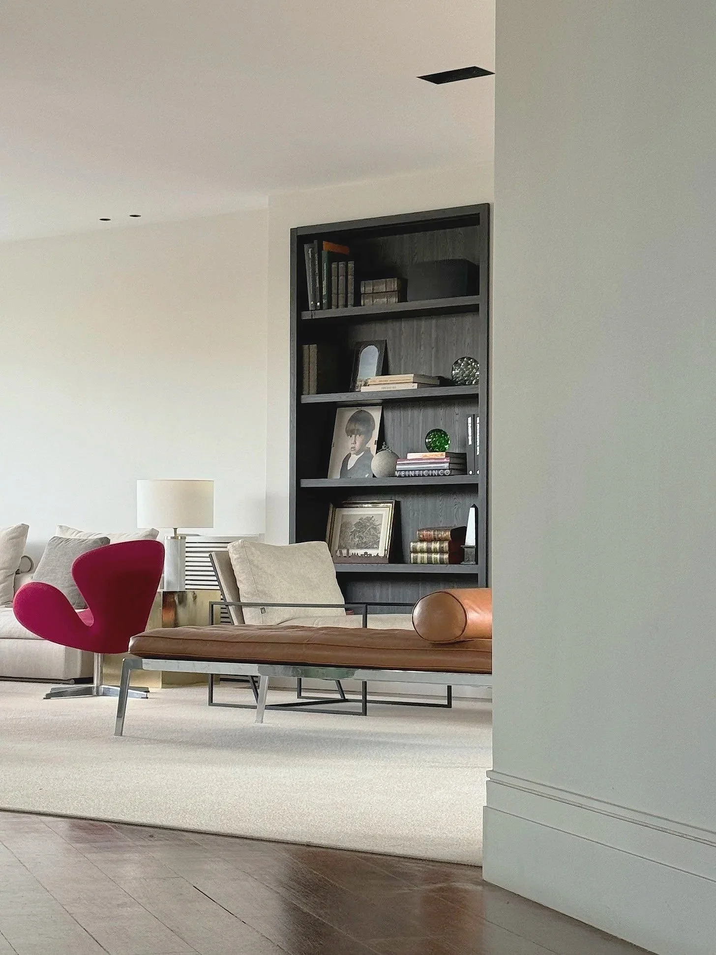 Living room with beige sofa, pink modern armchair, wooden bench with leather cushion, white lamp, and black bookshelf filled with books and decorative objects.